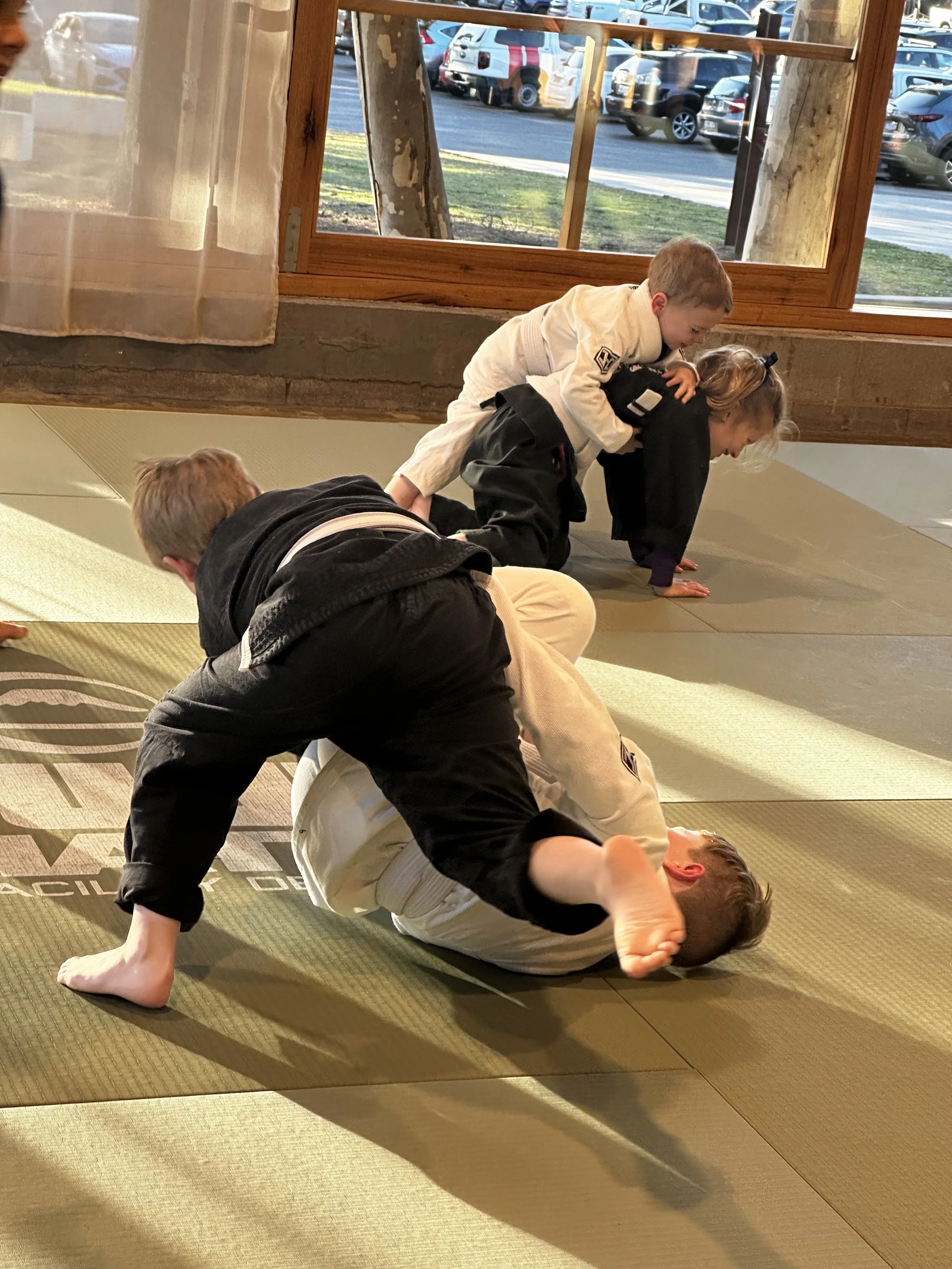Children practicing Brazilian Jiu-Jitsu on a mat indoors, with large windows showing parked cars outside.