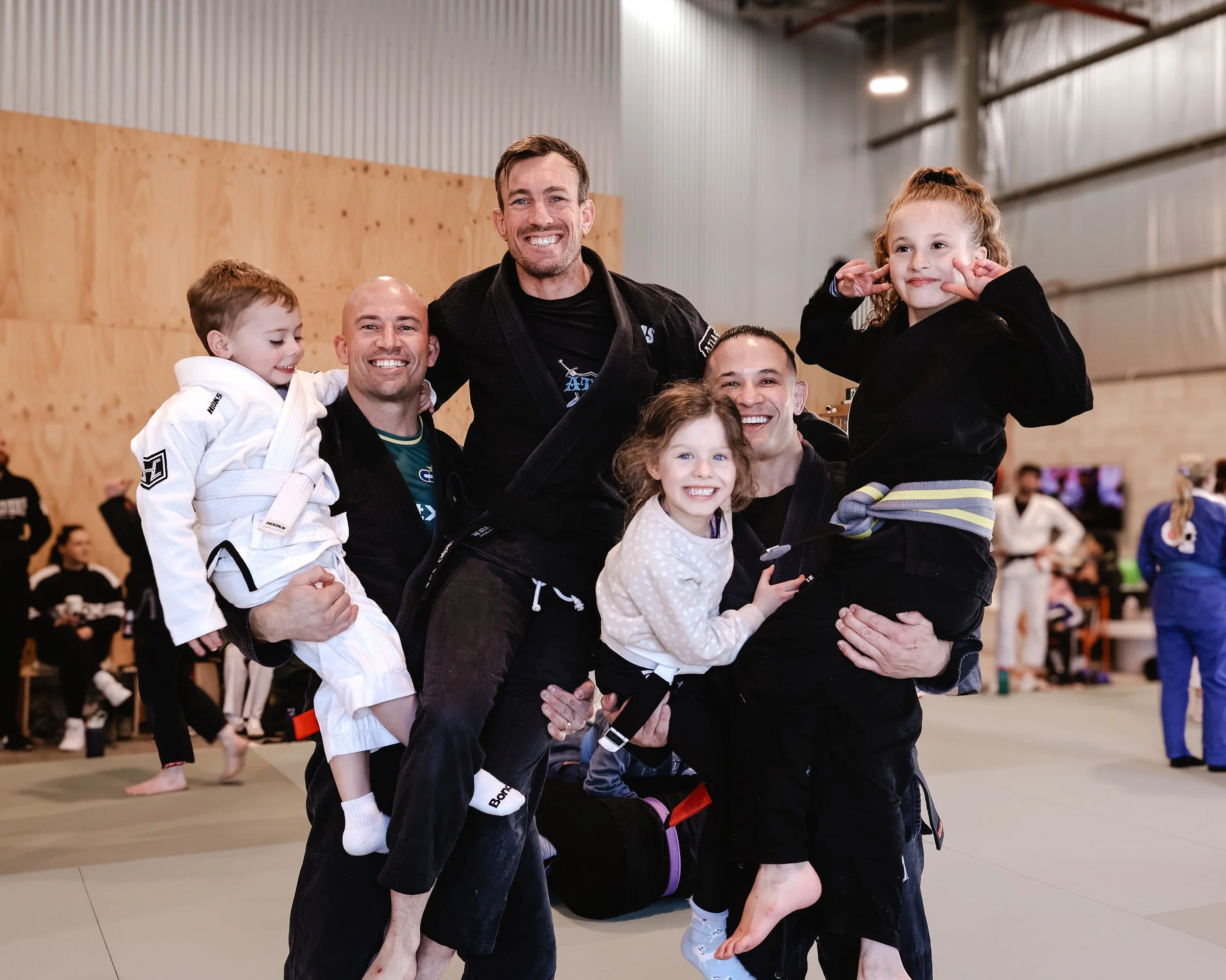 Group of people at a Brazilian Jiu-Jitsu training session, with children and adults, in an indoor gym. Some individuals are flexing or smiling.