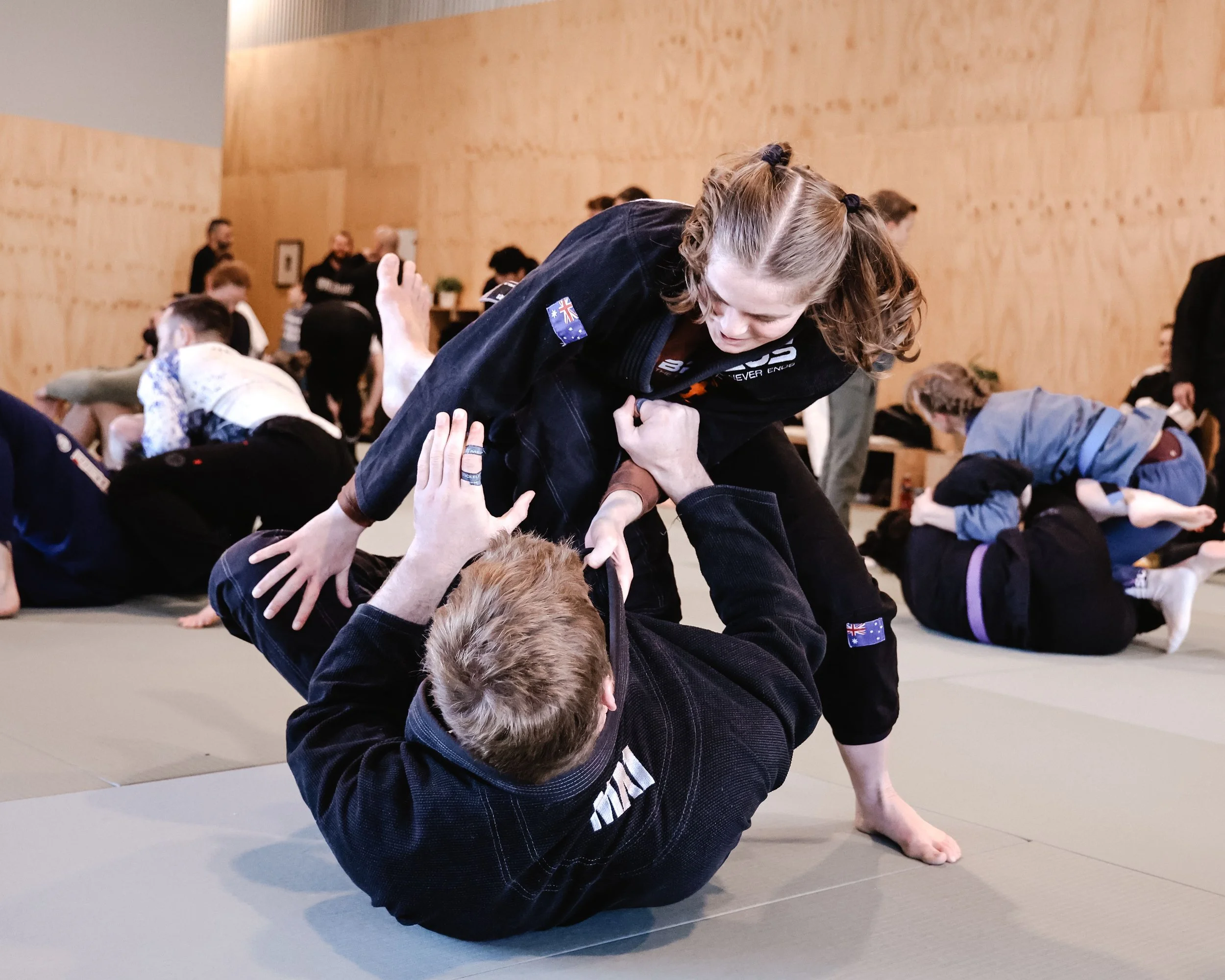 Two women practicing Brazilian Jiu-Jitsu on mats indoors, with one woman gripping the other's collar and sleeve while the other attempts to escape.