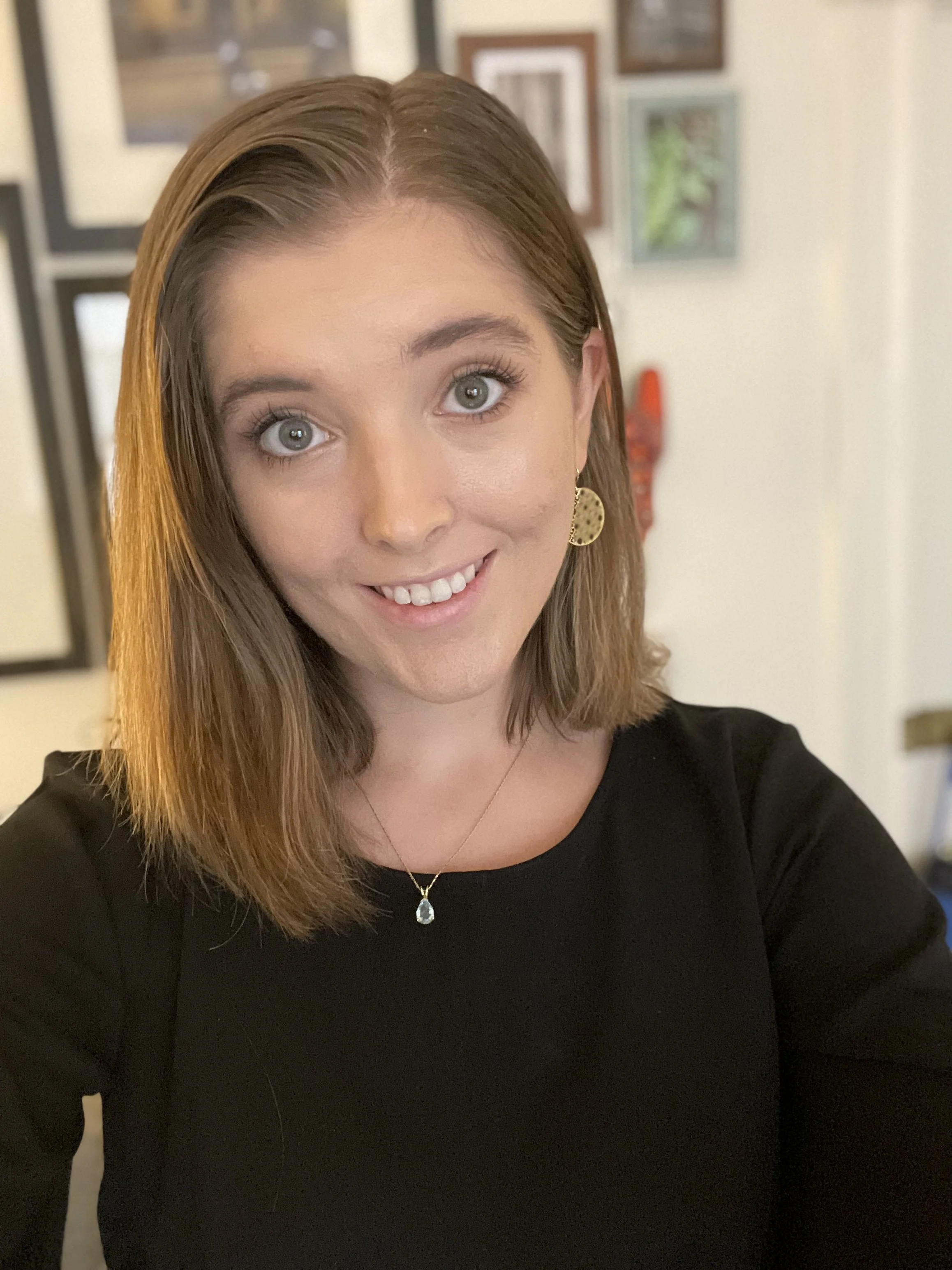 A woman with straight, shoulder-length brown hair taking a selfie. She is smiling, wearing a black top, gold dangle earrings, a necklace with a Teardrop gemstone pendant. The background is blurred with framed pictures on the wall.