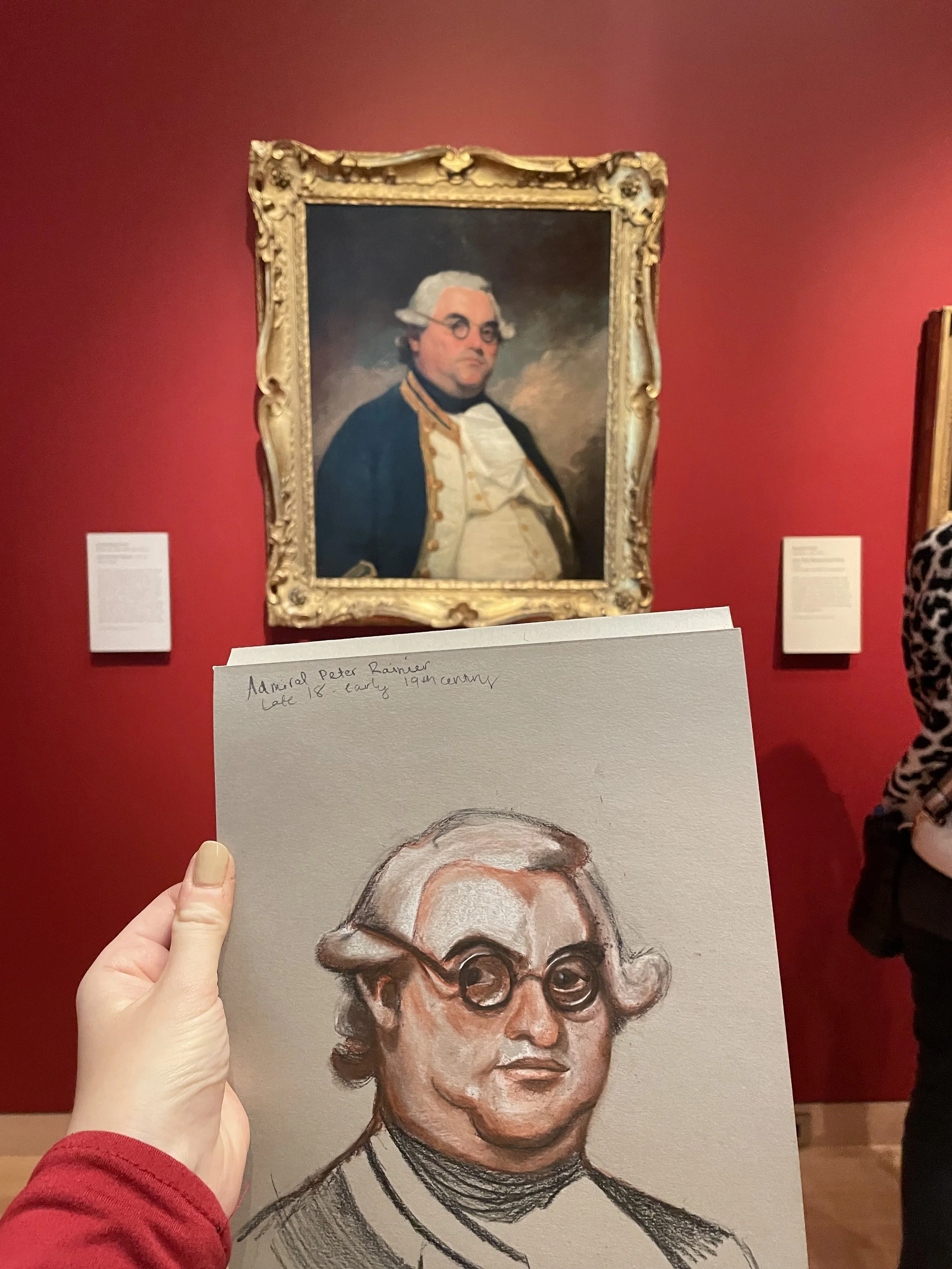 A person holding a drawing of a man wearing glasses, with a portrait of the same man in a gold ornate frame on a red wall in the background.