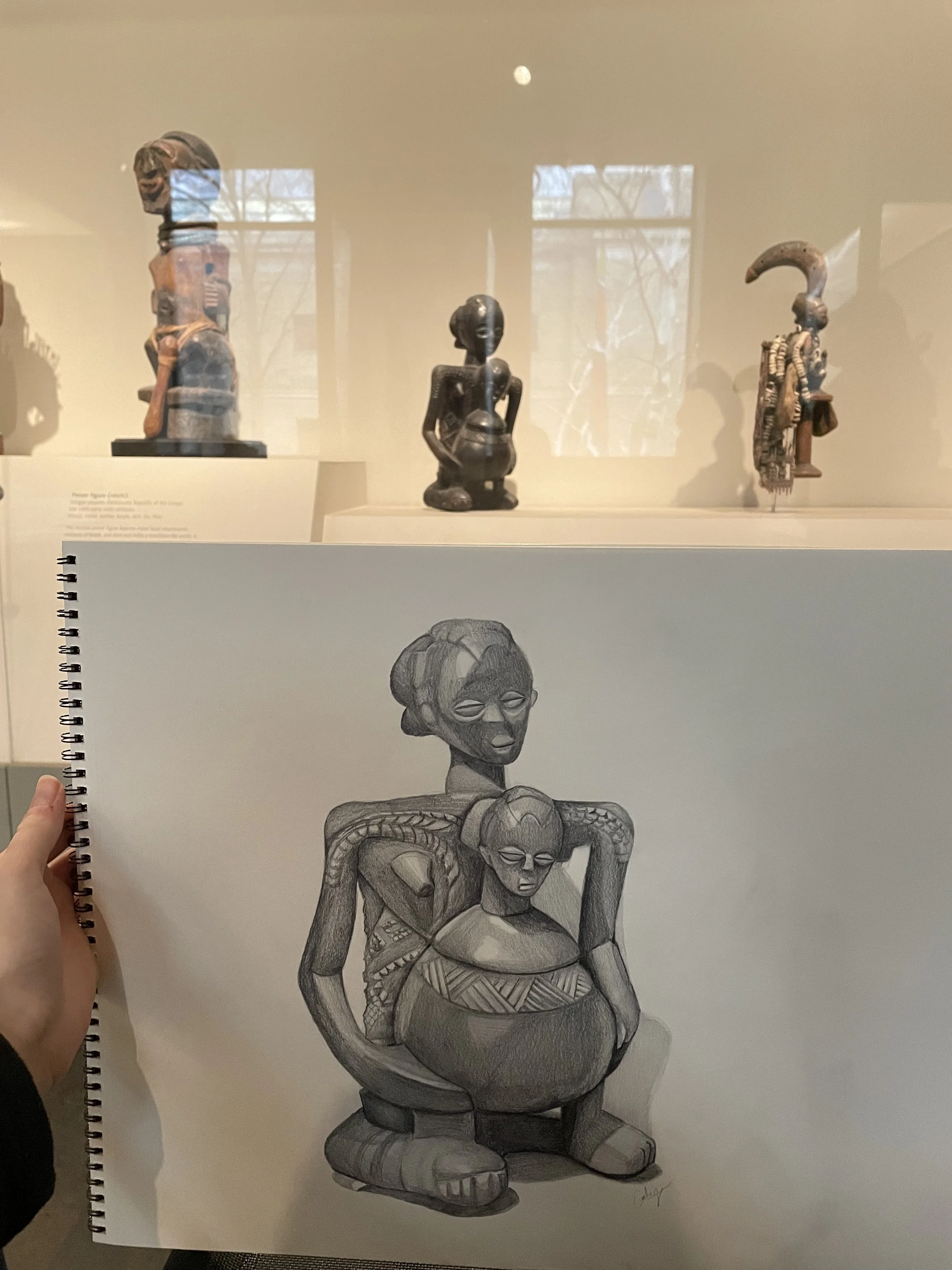 A black and white drawing of a tribal sculpture depicting two intertwined figures with detailed carvings, one larger and one smaller, with the larger figure sitting back and the smaller figure hugging its neck.