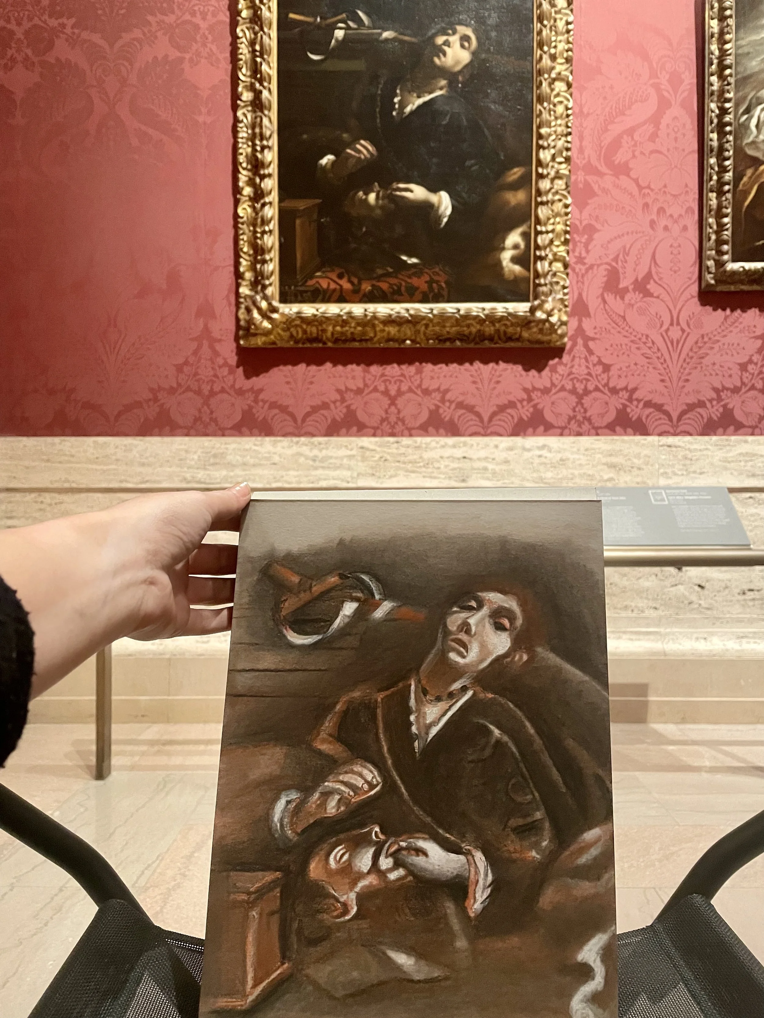 A person holding a printed replica of a painting depicting a woman with a snake around her neck, dressed in dark clothing, with an expressive face, in front of an original framed painting of the same woman on a pink wallpapered wall in an art gallery.