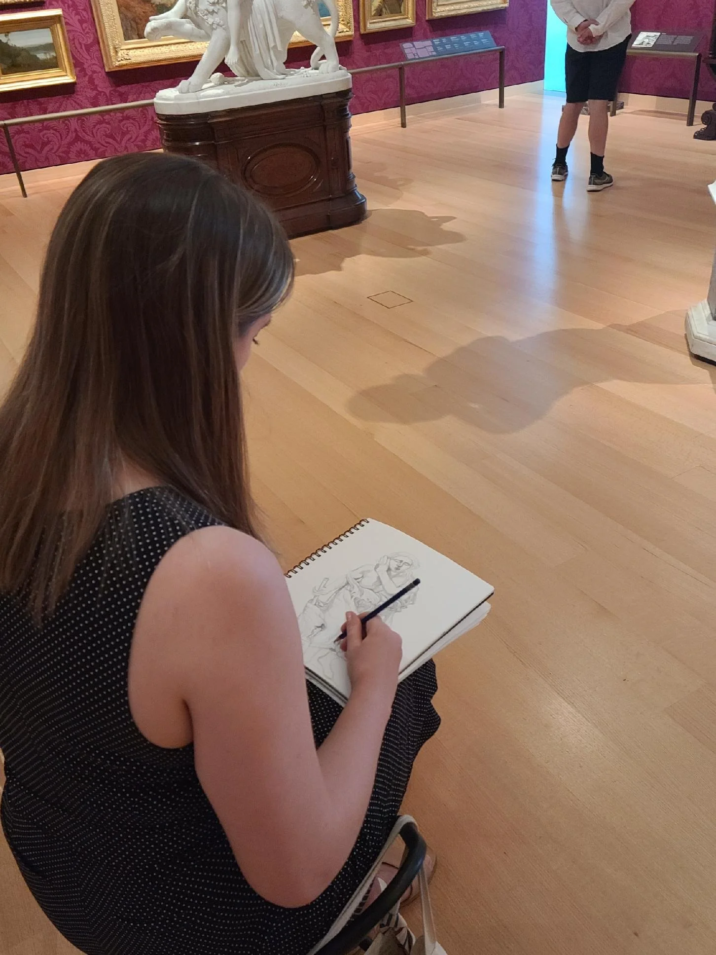 A girl with long brown hair sitting on a chair in a museum, drawing a sculpture of a human figure in her sketchbook. Other visitors are present in the background, viewing artworks displayed on the wall.
