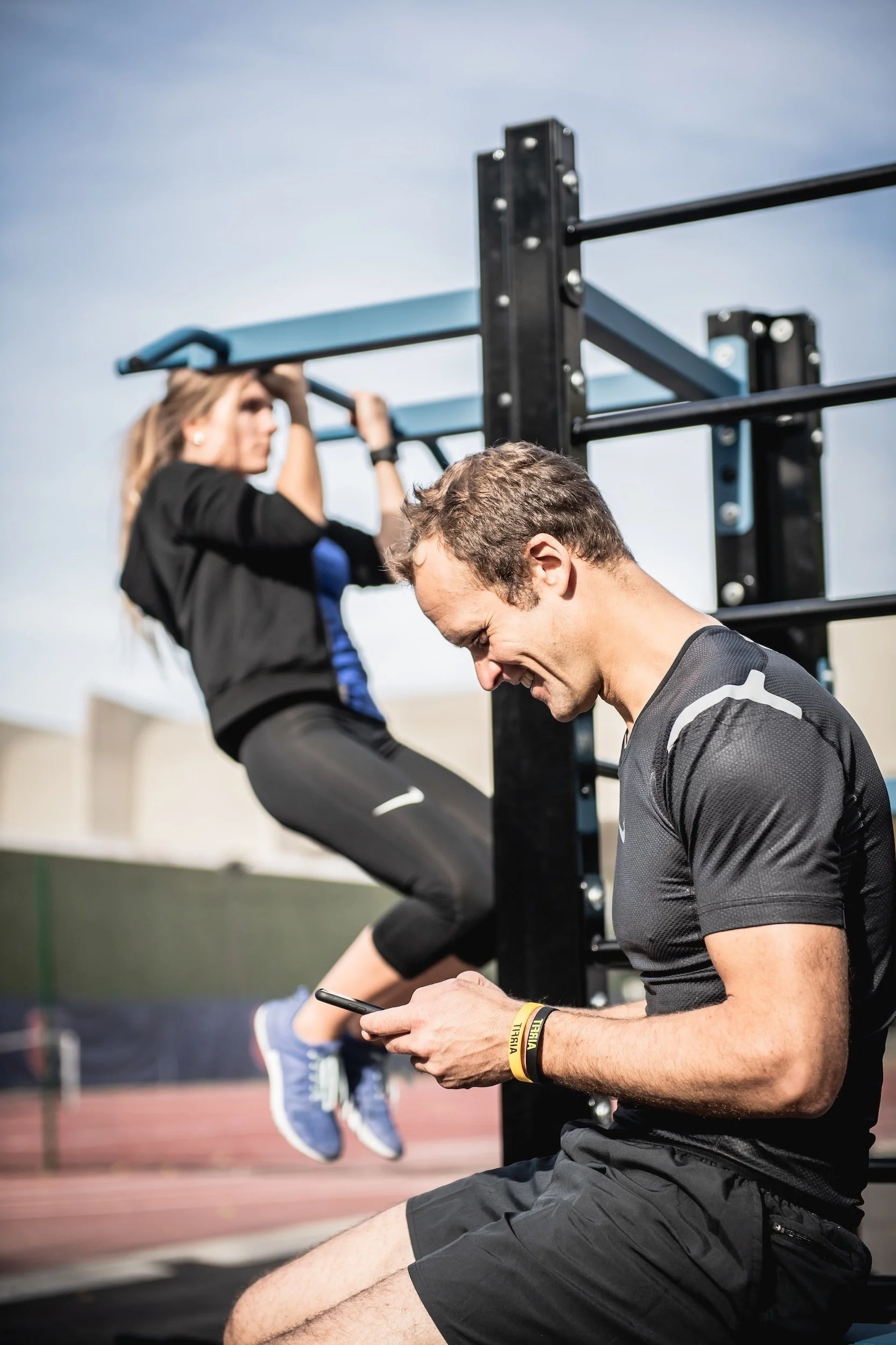 A man sits on a bench at an outdoor gym, looking at his phone while a woman exercises on a pull-up bar in the background.