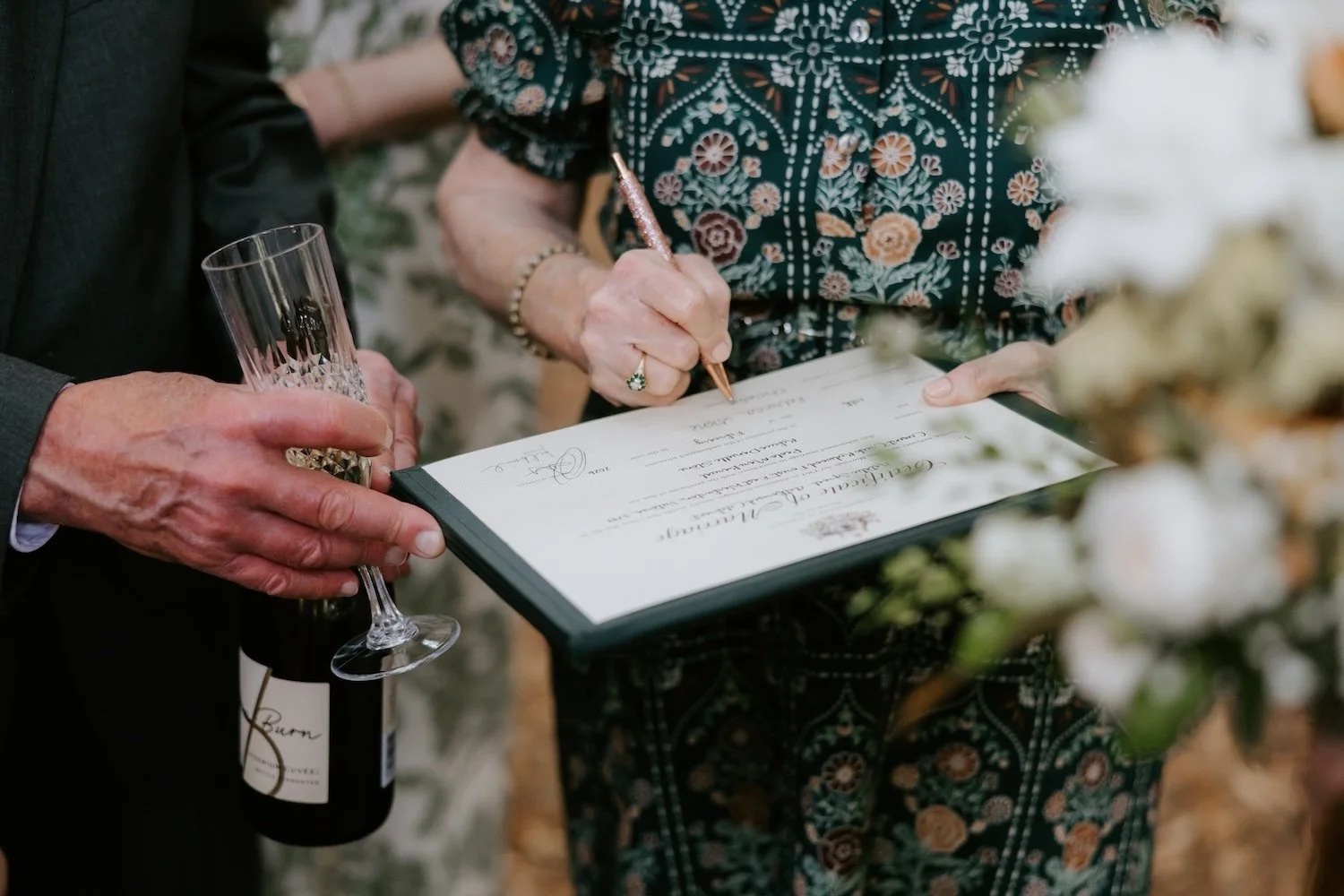 A close up image of a bride's Mum signing her marriage certificate as a witness. Also in frame is a bottle of champagne and a glass.