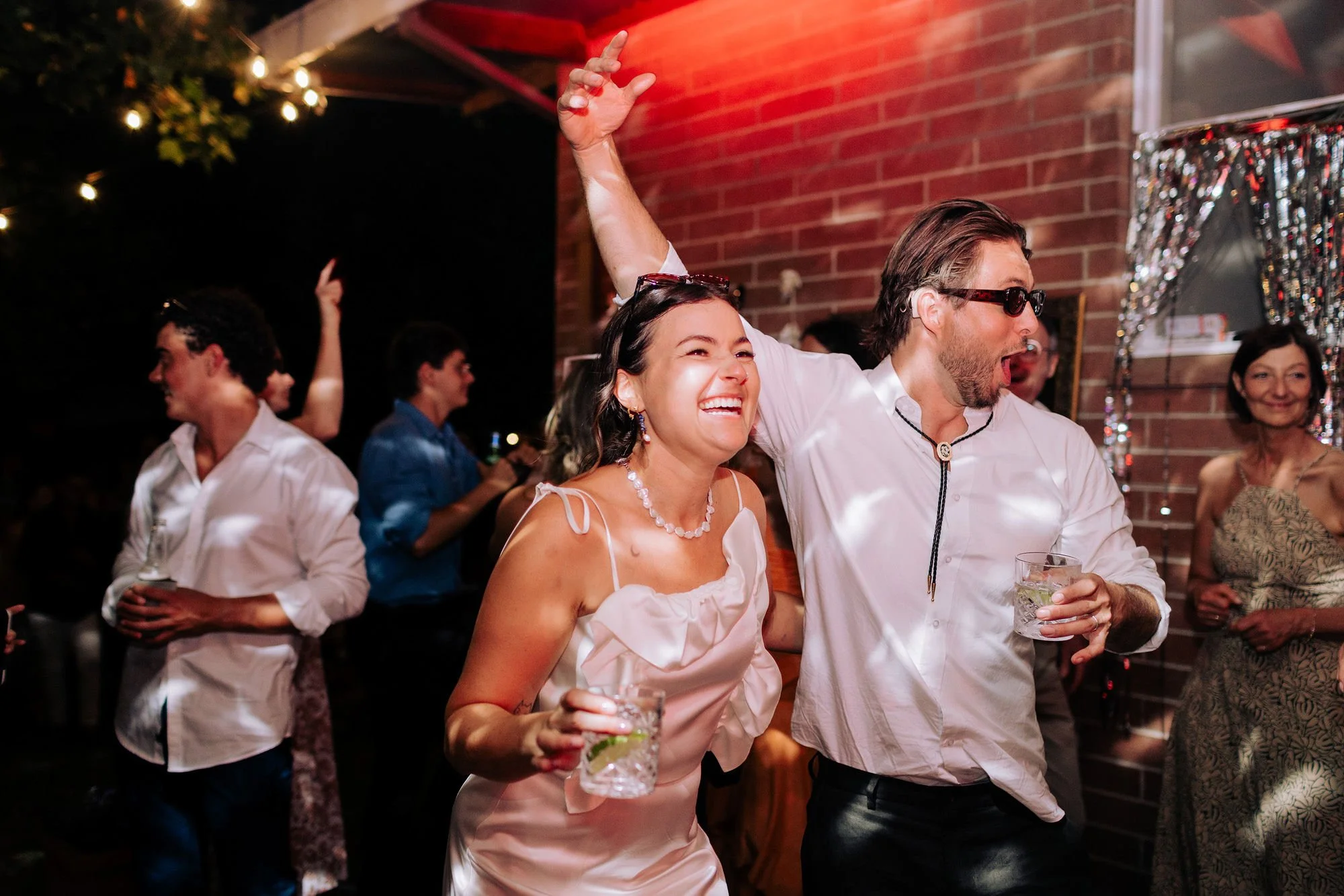 A bride and groom wearing sunglasses and holding gin and tonics are having the best time on the dance floor together.