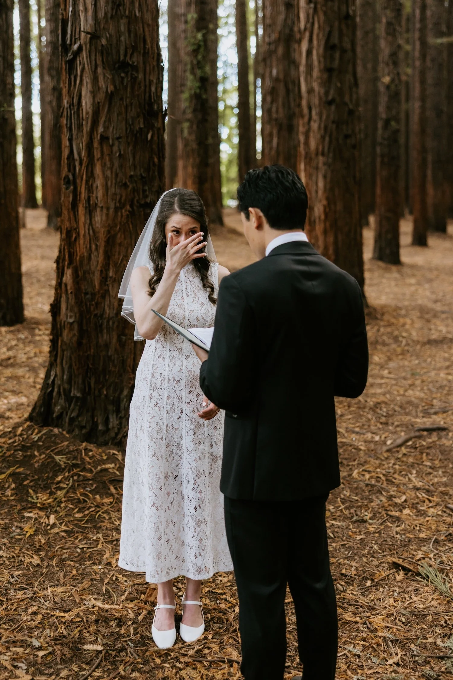 An emotional bride in a white lace dress wipes away tears as her groom reads his vows to her in the Redwood Forest at Warburton.