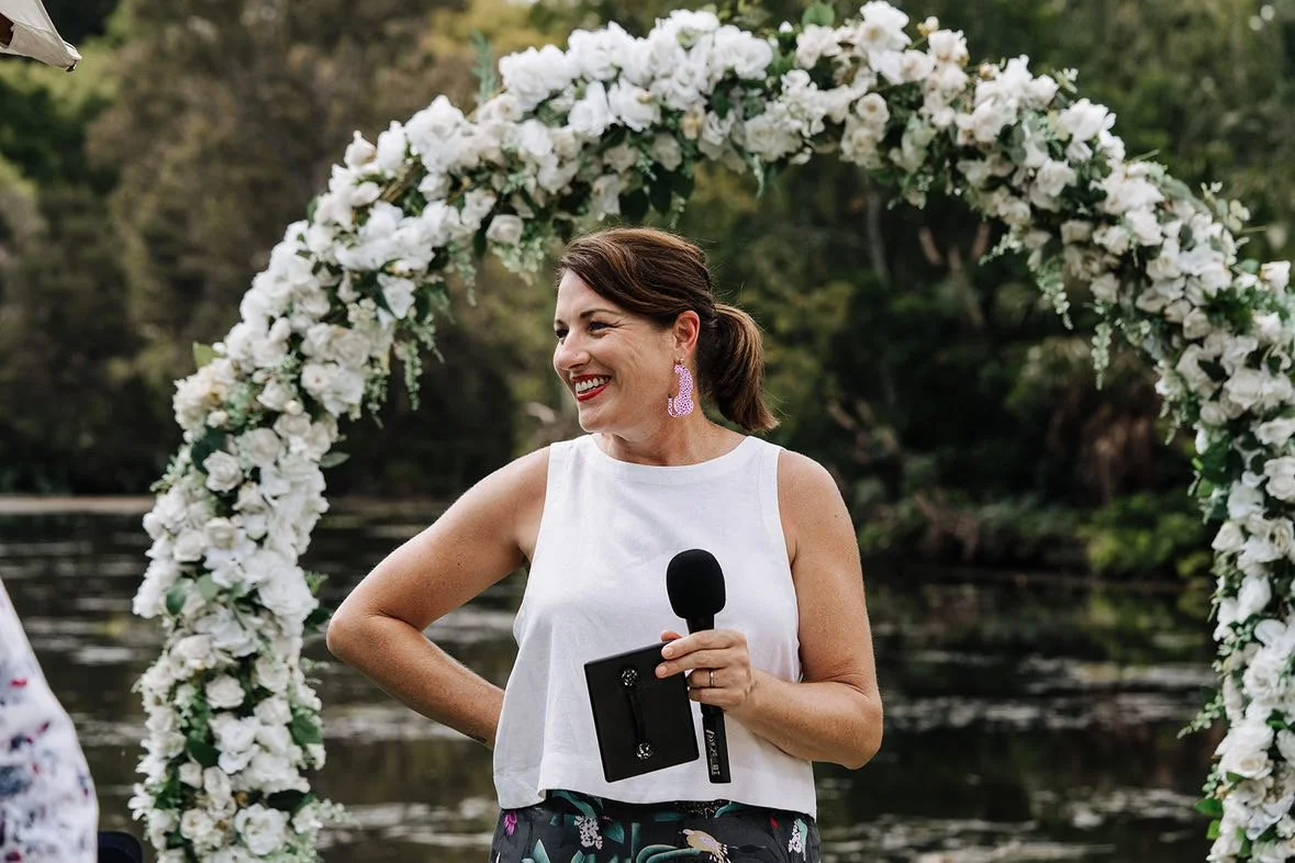 A candid shot of Nat Sproal holding a microphone and Kindle smiling at someone to the left of her. Behind her is a lake and a circular arbour adorned with white flowers