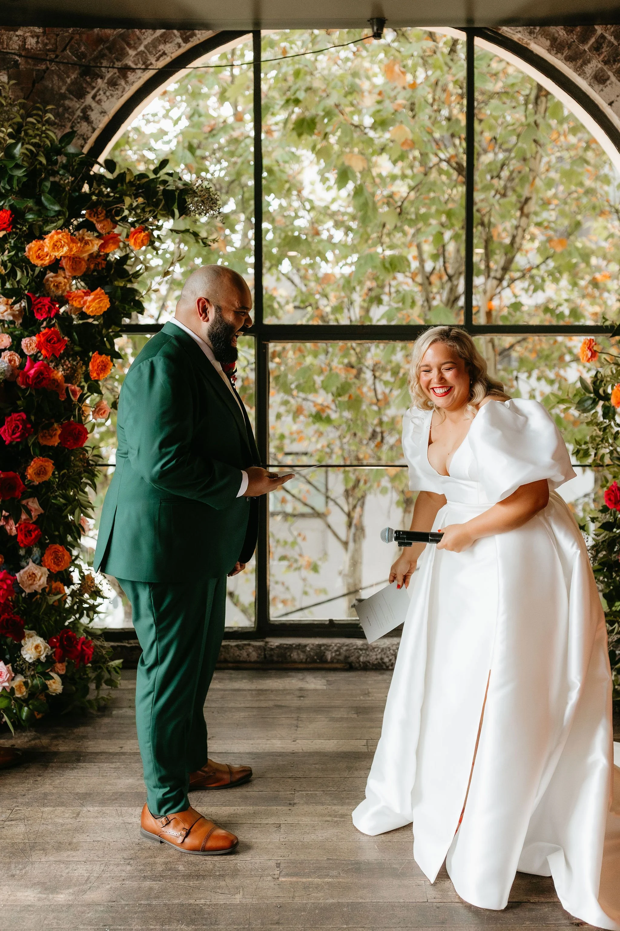 Profile shot of a groom in a green suit and his bride in a white gown laughing as they read their vows to each other. In the background the arch window is adorned with red and orange roses.