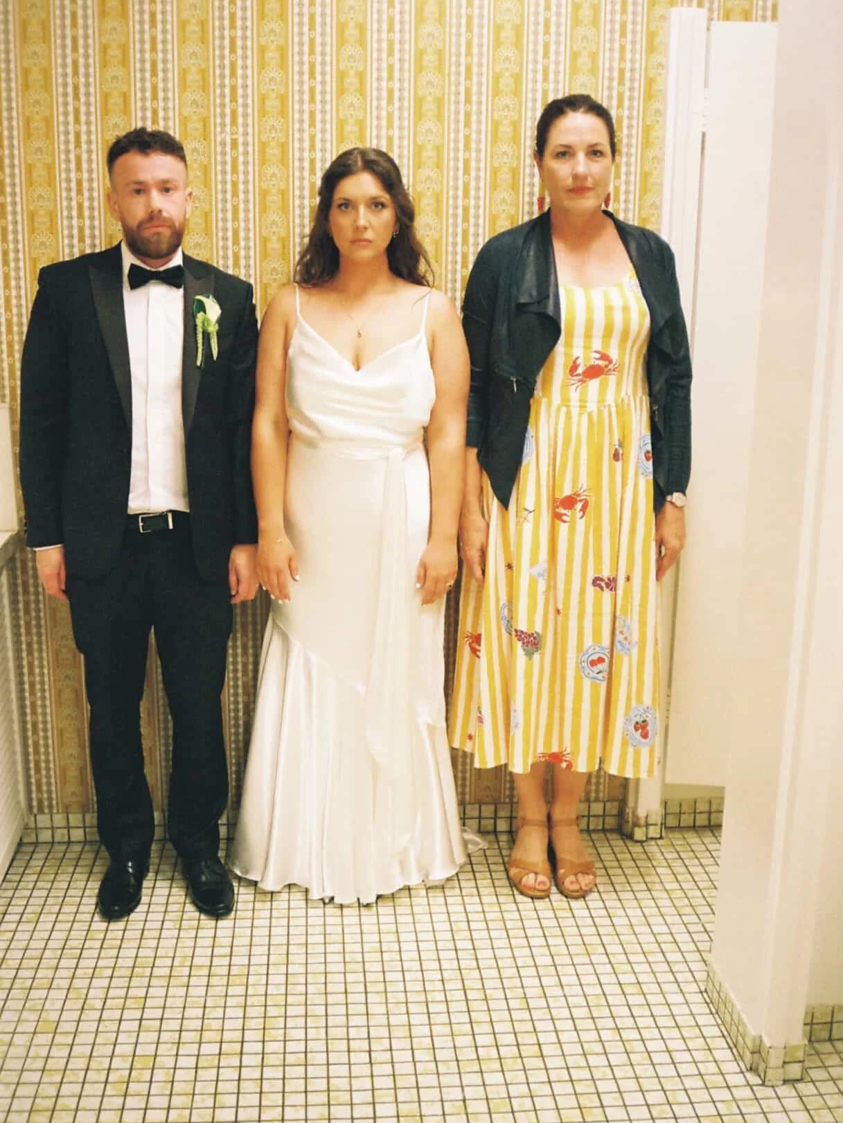 Melbourne celebrant Nat Sproal is pictured with a bride and groom standing full face on, arms straight by sides with serious faces against retro yellow wallpaper, Wes Anderson style