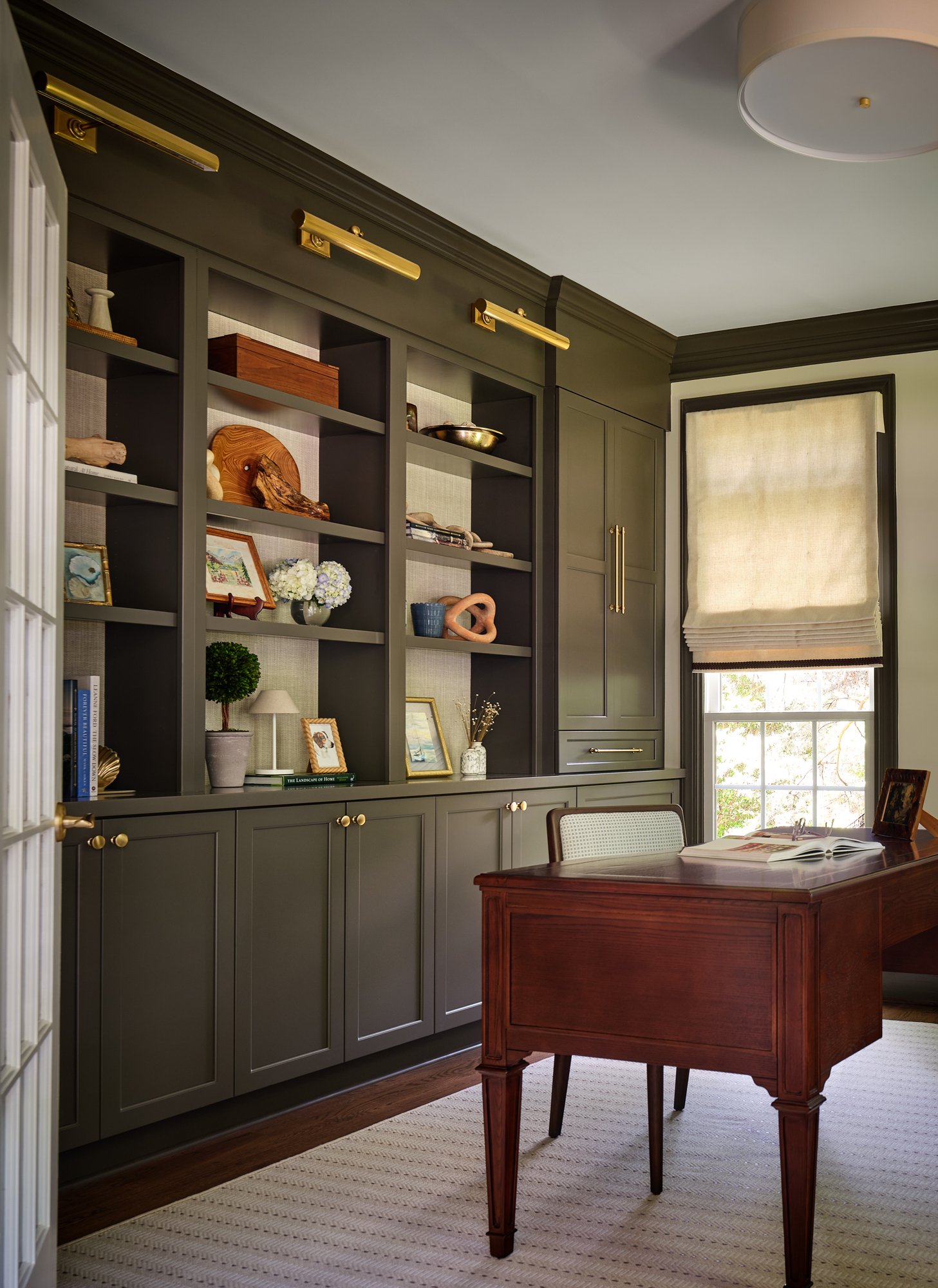 Interior of a room with a large gray bookshelf stocked with decorative items, framed photos, and books, a window with a beige shade, a wooden desk with an open book, and a beige and white striped rug on a hardwood floor.