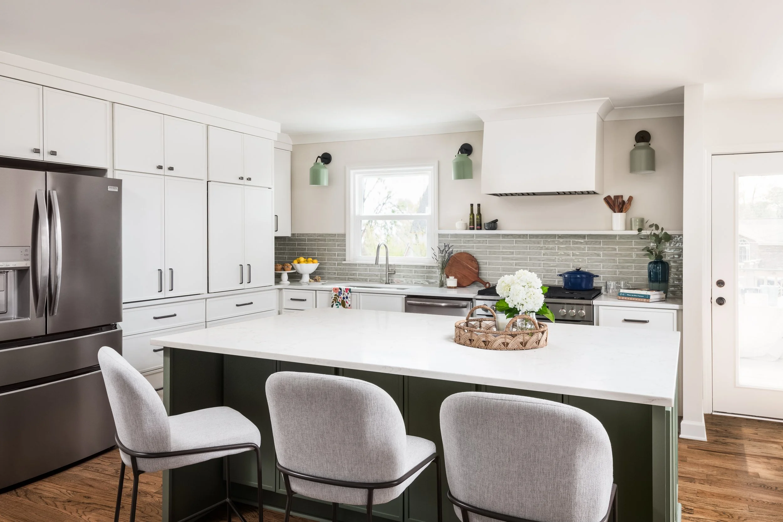 Modern kitchen with white cabinets, stainless steel refrigerator, green island with dining chairs, brick backsplash, window, and various decor items.
