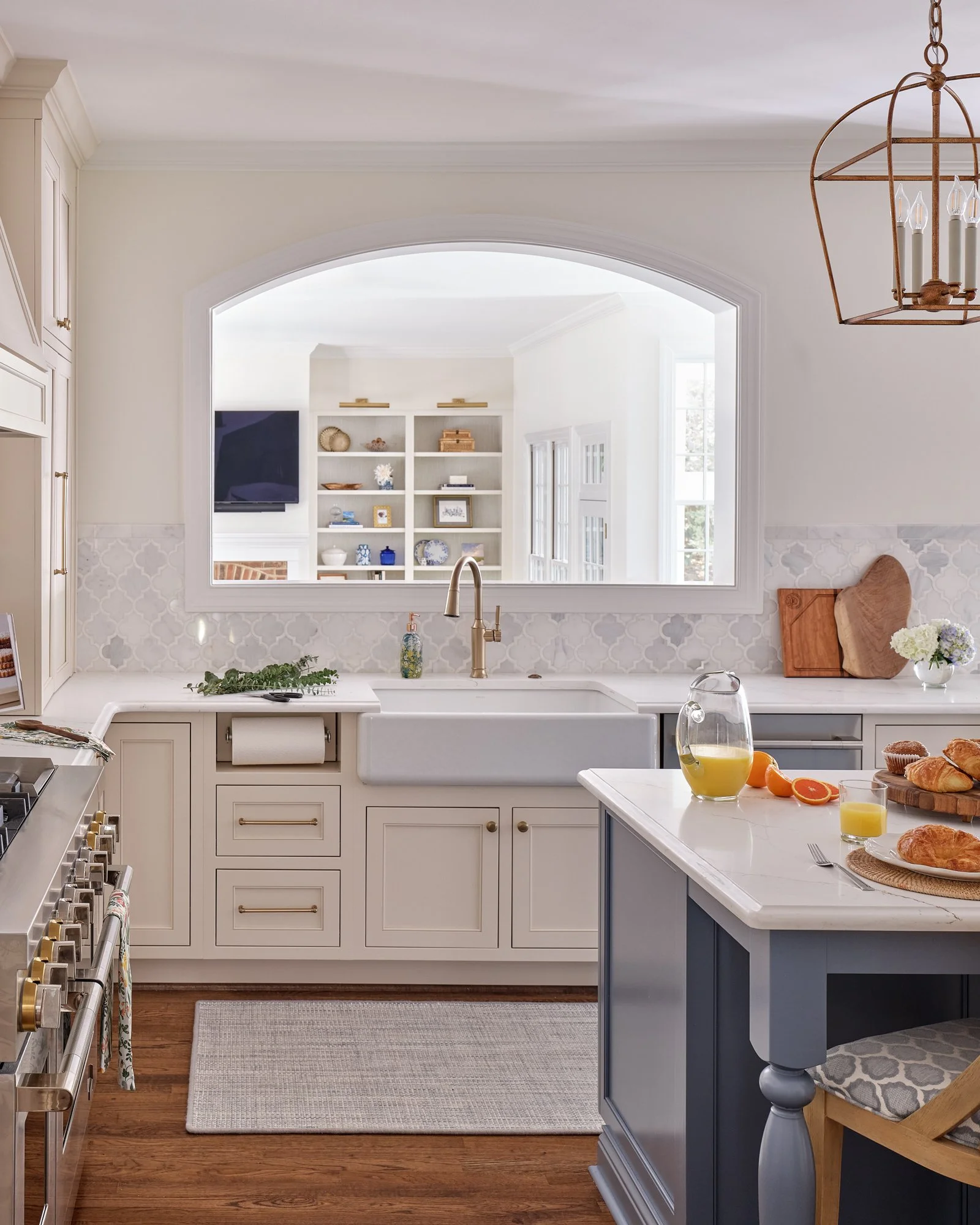 A kitchen interior view featuring a white farmhouse sink, a white island with breakfast items, and built-in shelves with decorative items in the background. The kitchen has a warm hardwood floor, a gray-blue lower cabinet, and a chandelier hanging from the ceiling.