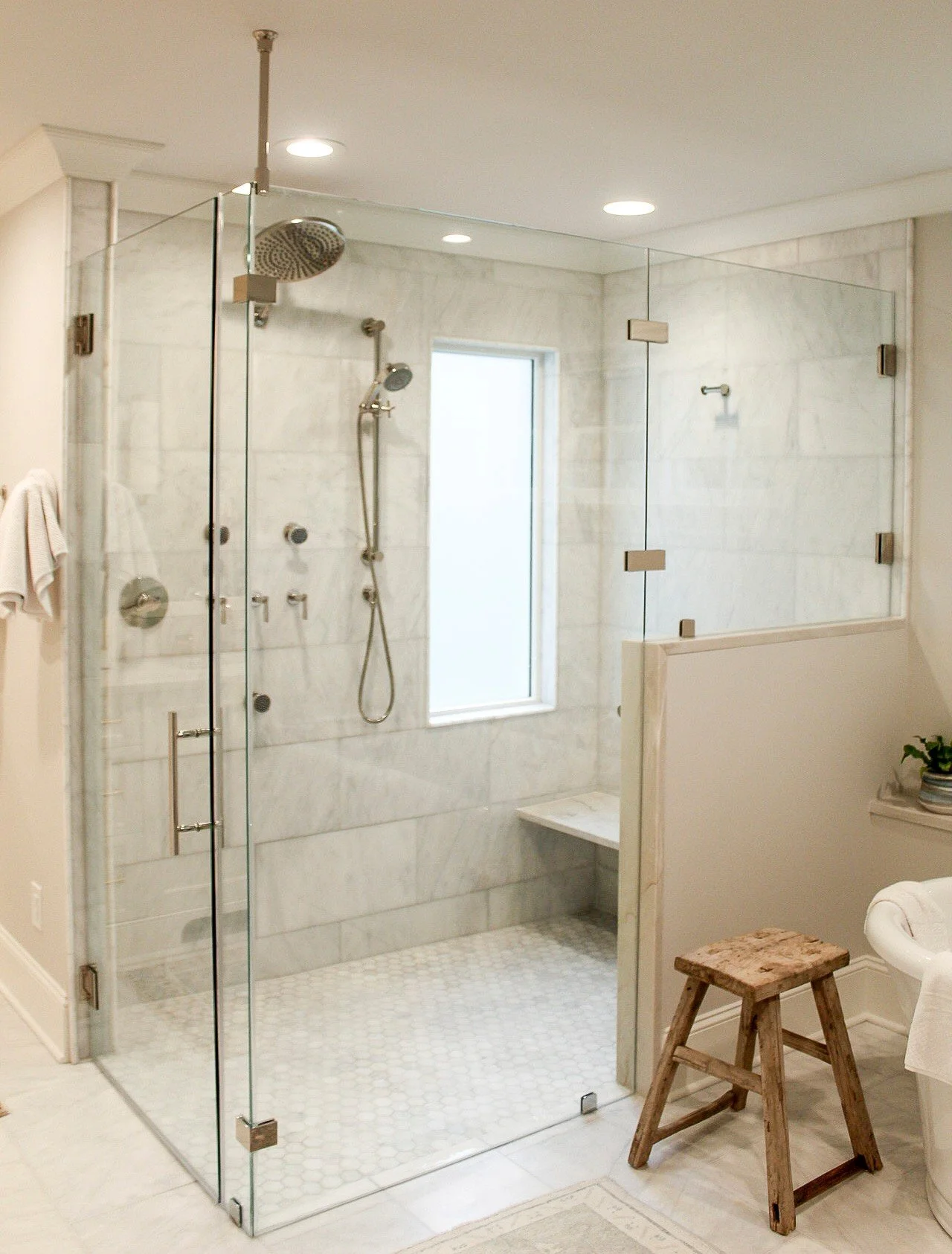 This South Charlotte zero-entry shower features a seamless design paired with a statement shower system, including an oversized rain head, handheld, and body sprayers for a true spa-at-home experience. Clean marble walls, a built-in bench, and framel