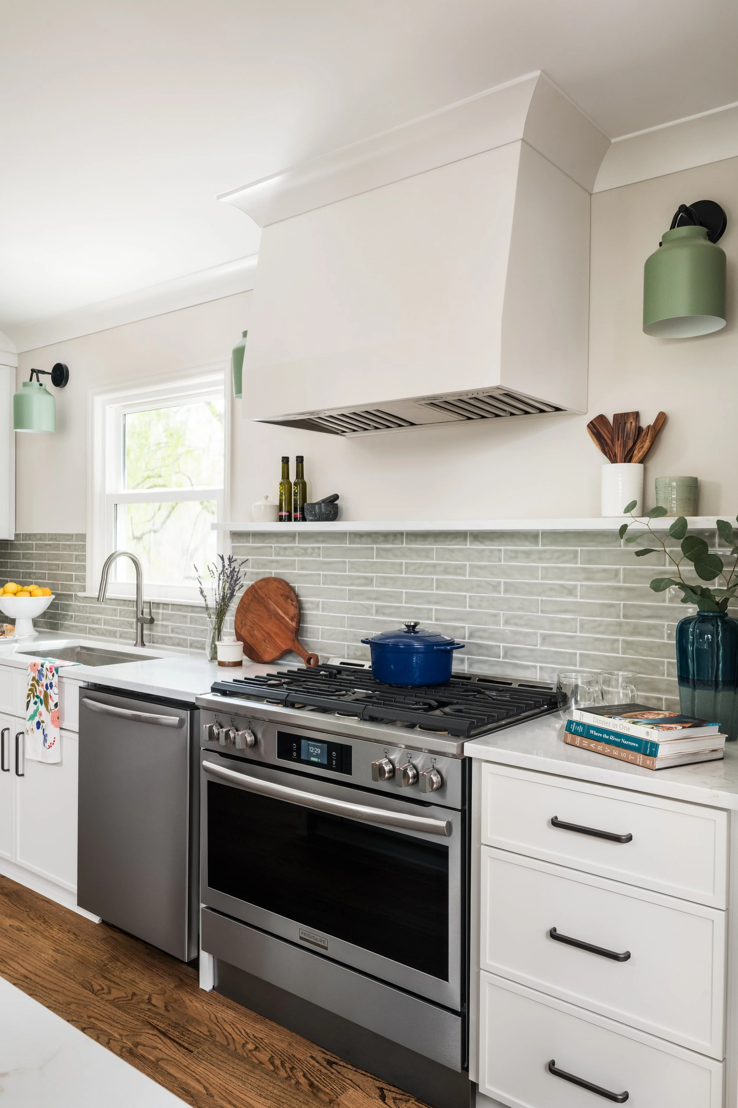 A modern kitchen with a stainless steel stove, a white countertop, white cabinets, a window with a view of greenery, and kitchen decorations including a bowl of lemons, a cutting board, and books on the counter.