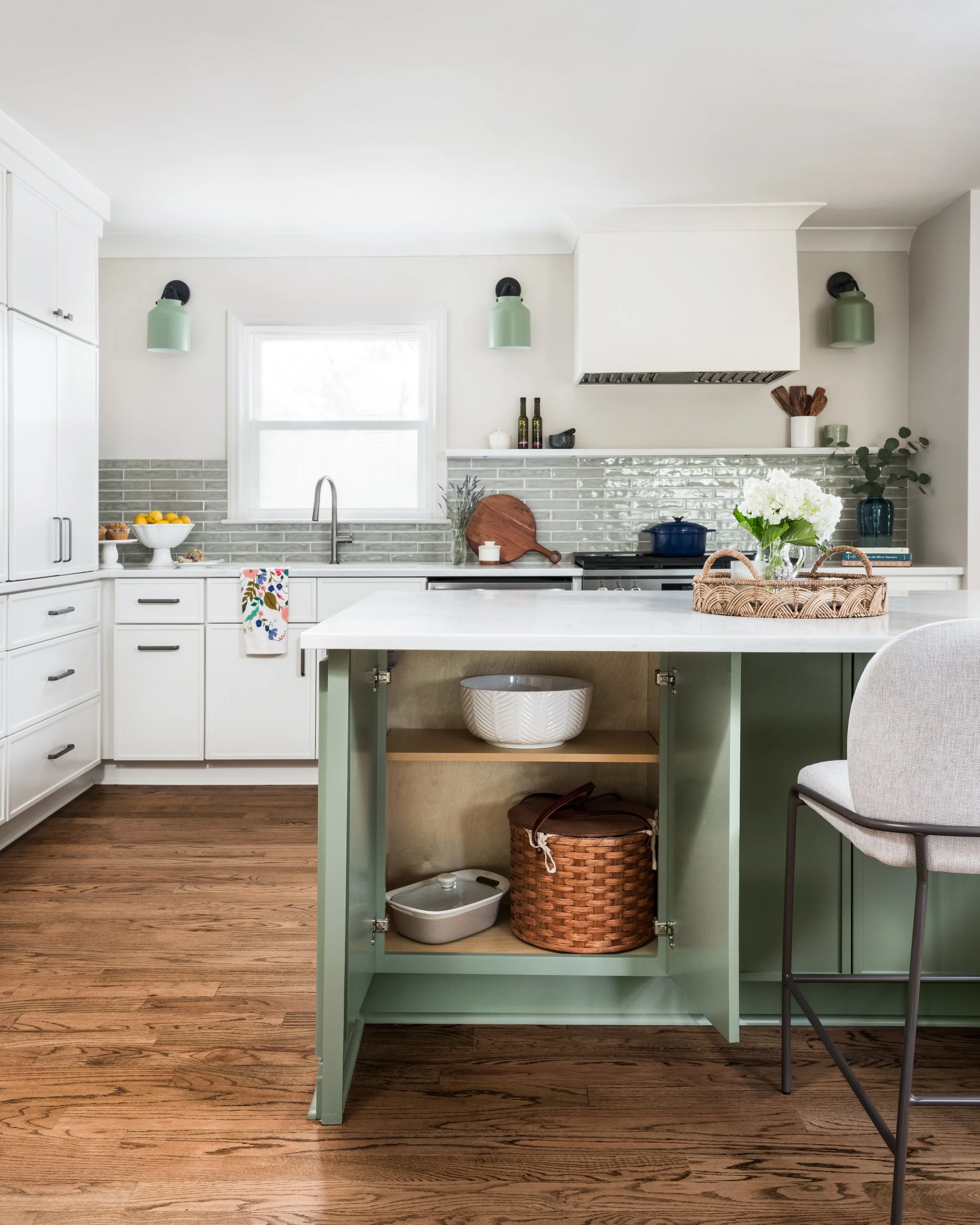 A modern kitchen with white cabinetry, a kitchen island with a green base, a white countertop, light green wall sconces, and wooden flooring. The kitchen features a window, a bowl of lemons, and various decorative items.