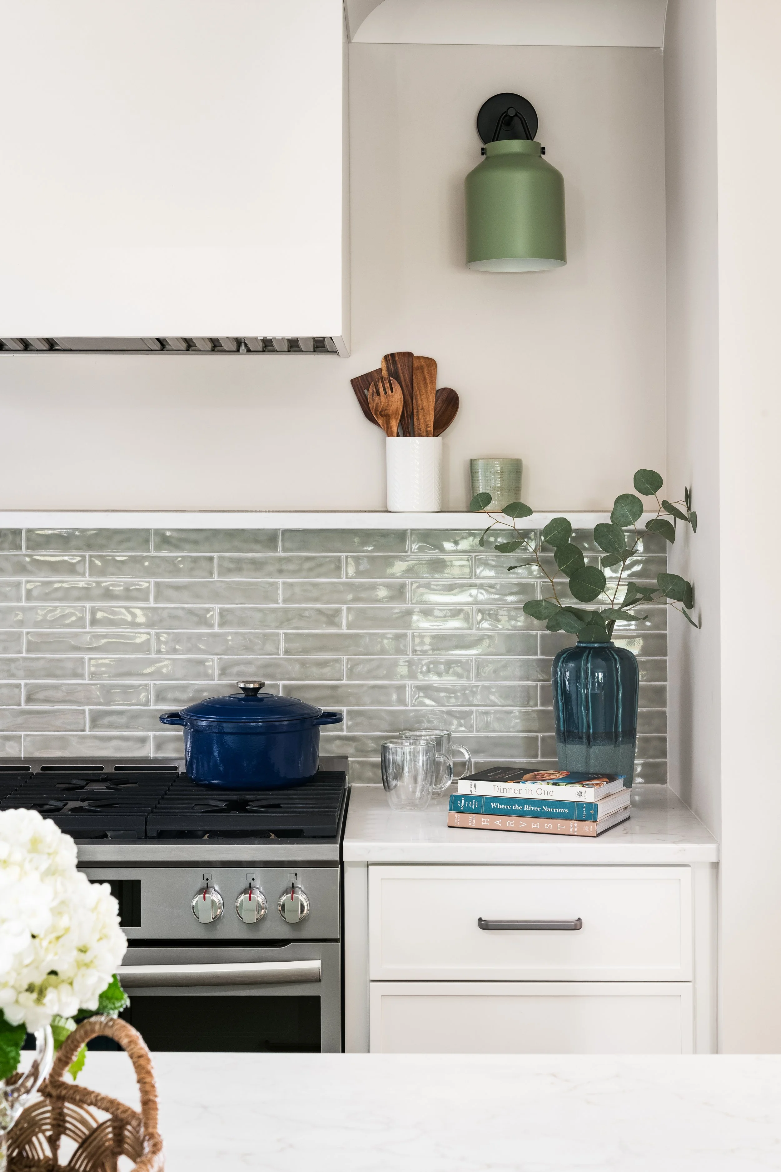 Kitchen countertop with a stove, a blue pot, books, cups, a vase with green leafy branches, and kitchen utensils in a white container.