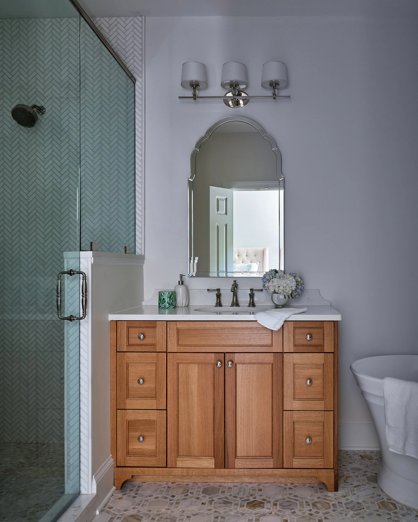 A bathroom vanity with a wooden cabinet and a white countertop. There are decorative soap dispensers and a bouquet of white flowers on the counter. A large mirror with a decorative frame hangs above the vanity. On the left side, there is a shower are