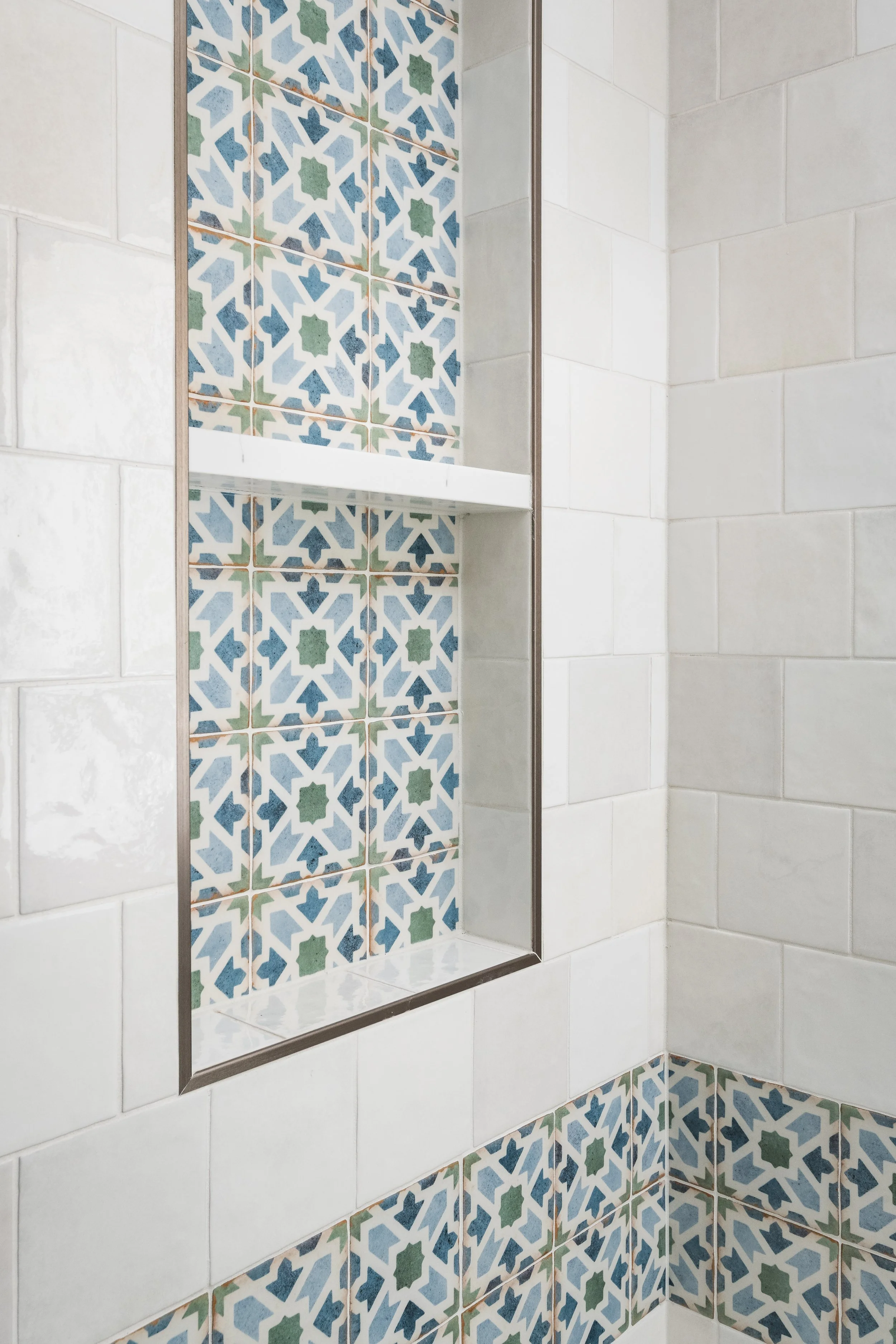 Close-up of a shower with decorative and plain tiles. The decorative tiles have an intricate geometric pattern in blue, green, and white, while the plain tiles are white. There is a glass shower door and a white shelf inside the shower.