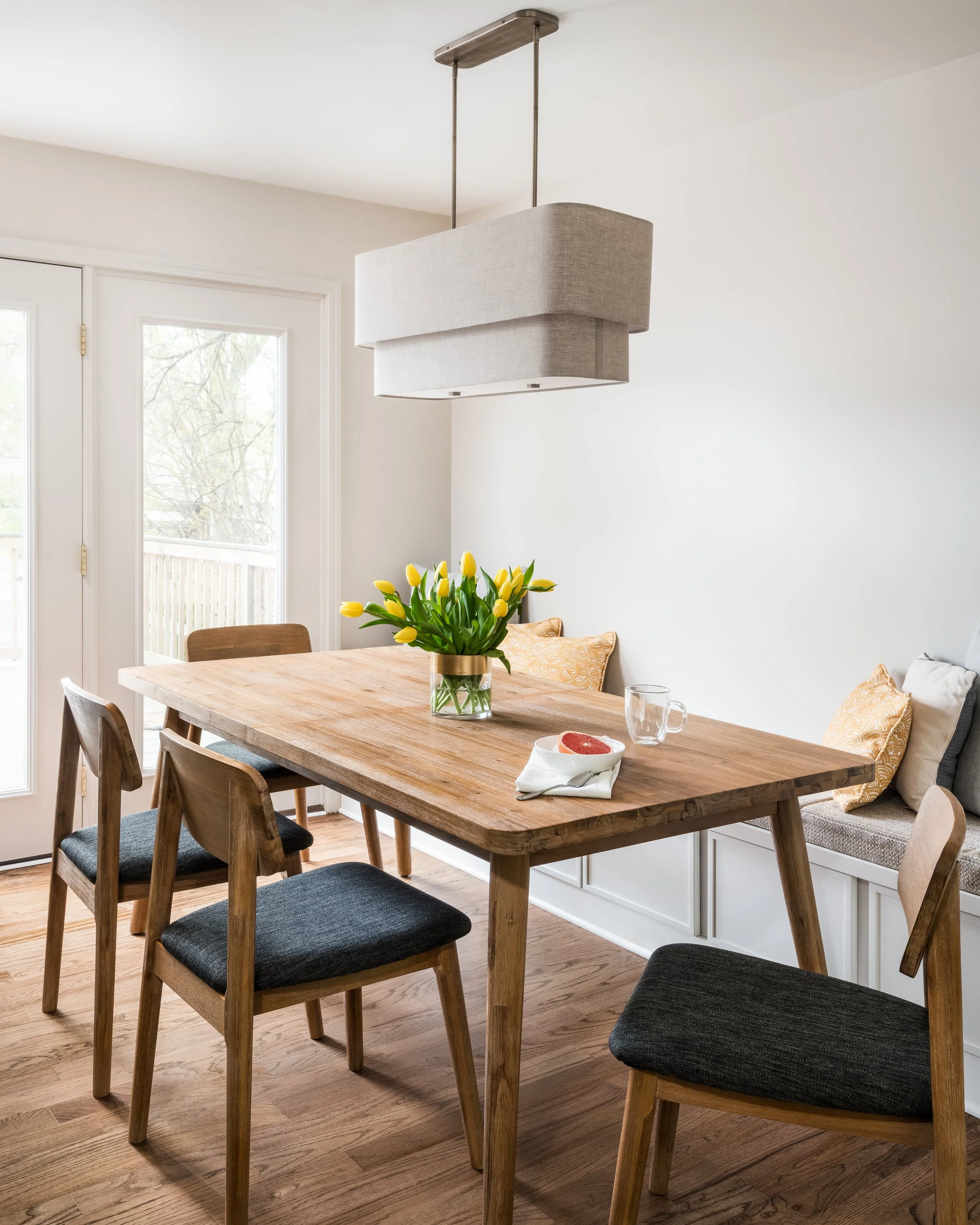 A dining room with a wooden table, six chairs with black cushions, and a hanging modern light fixture. On the table, there is a vase with yellow tulips, a grapefruit, a glass mug, and a white napkin with a spoon.
