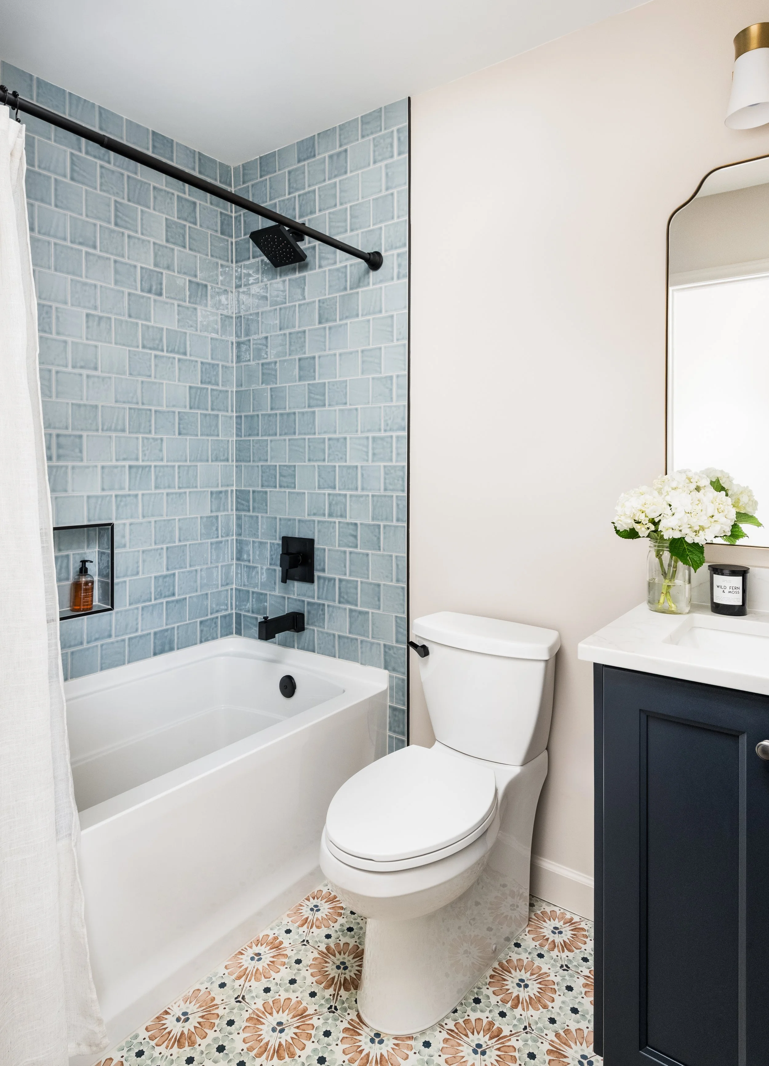 A bathroom with blue tiled shower area, black fixtures, a white bathtub, a toilet, a dark blue bathroom vanity, a mirror, a vase with white flowers, and patterned tile flooring.