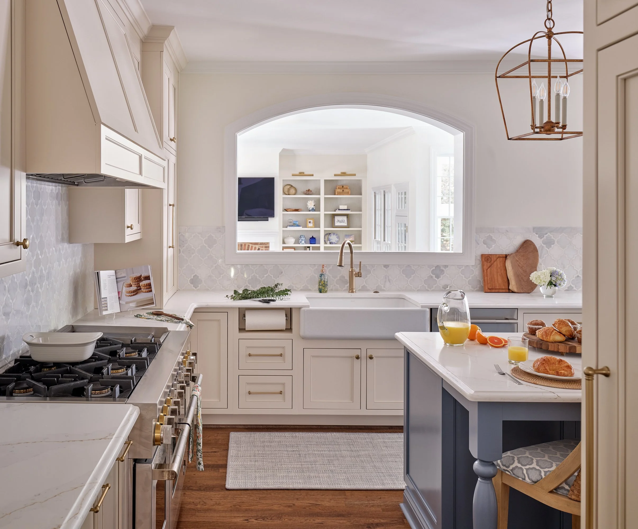 A kitchen with white cabinets, a white island with a blue base, and a gas stove. There is a large arched window above the sink, a wooden chair, and a breakfast table with croissants, orange juice, and flowers on it. A chandelier hangs from the ceilin