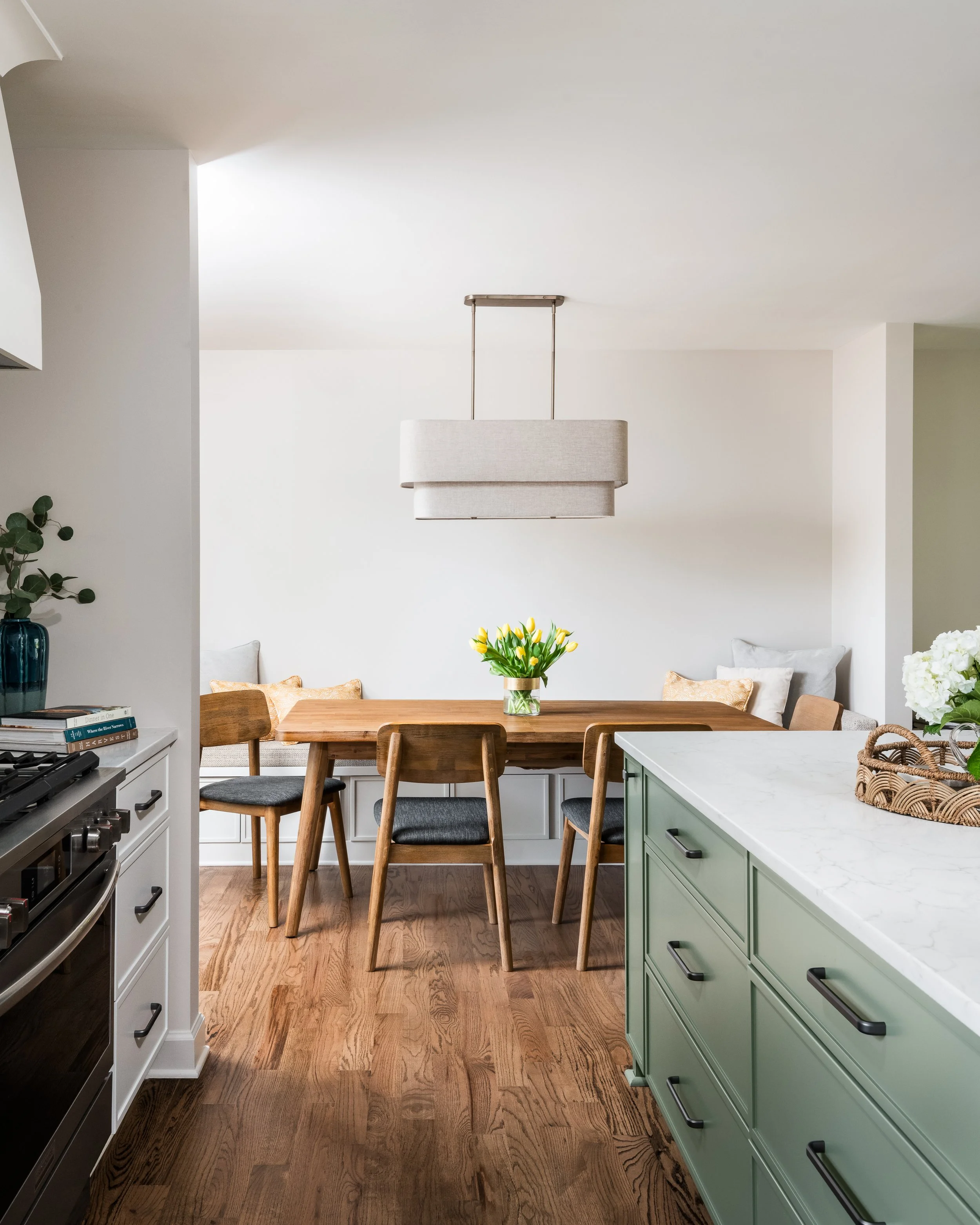 Modern kitchen and dining area with a wooden table, chairs, a pendant light, a green cabinet, and white walls decorated with books, plants, and flowers.
