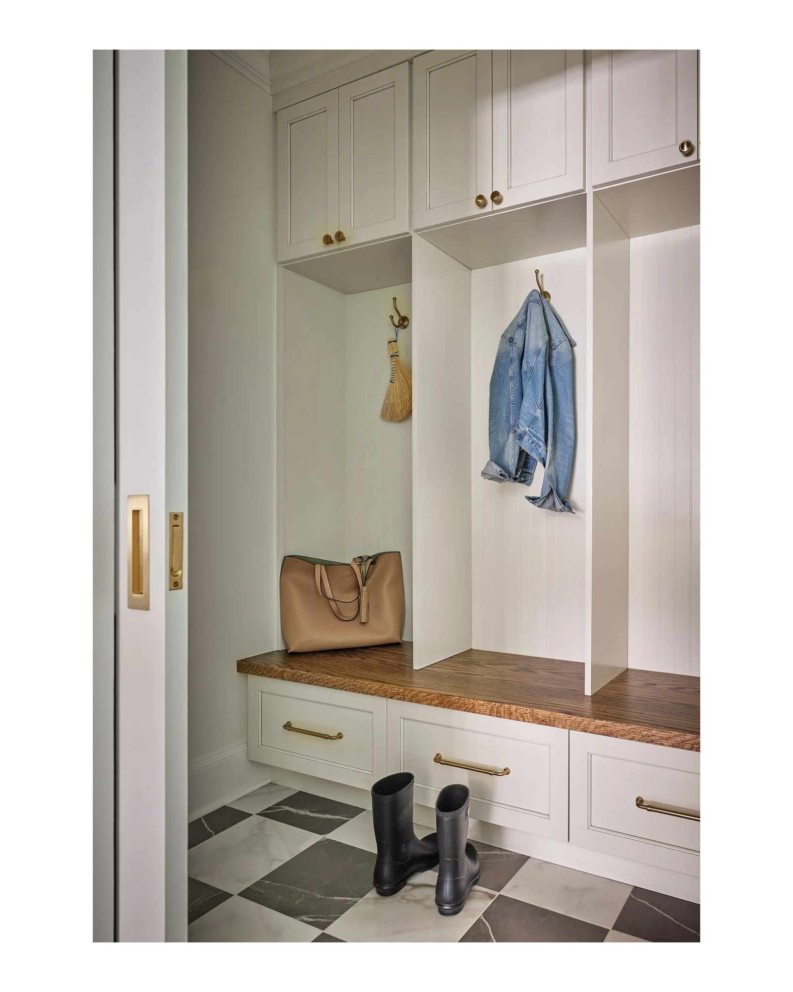 A foyer with white wall paneling, gold hardware, a wooden shelf, a beige tote bag, black rain boots, and black and white tiled flooring.