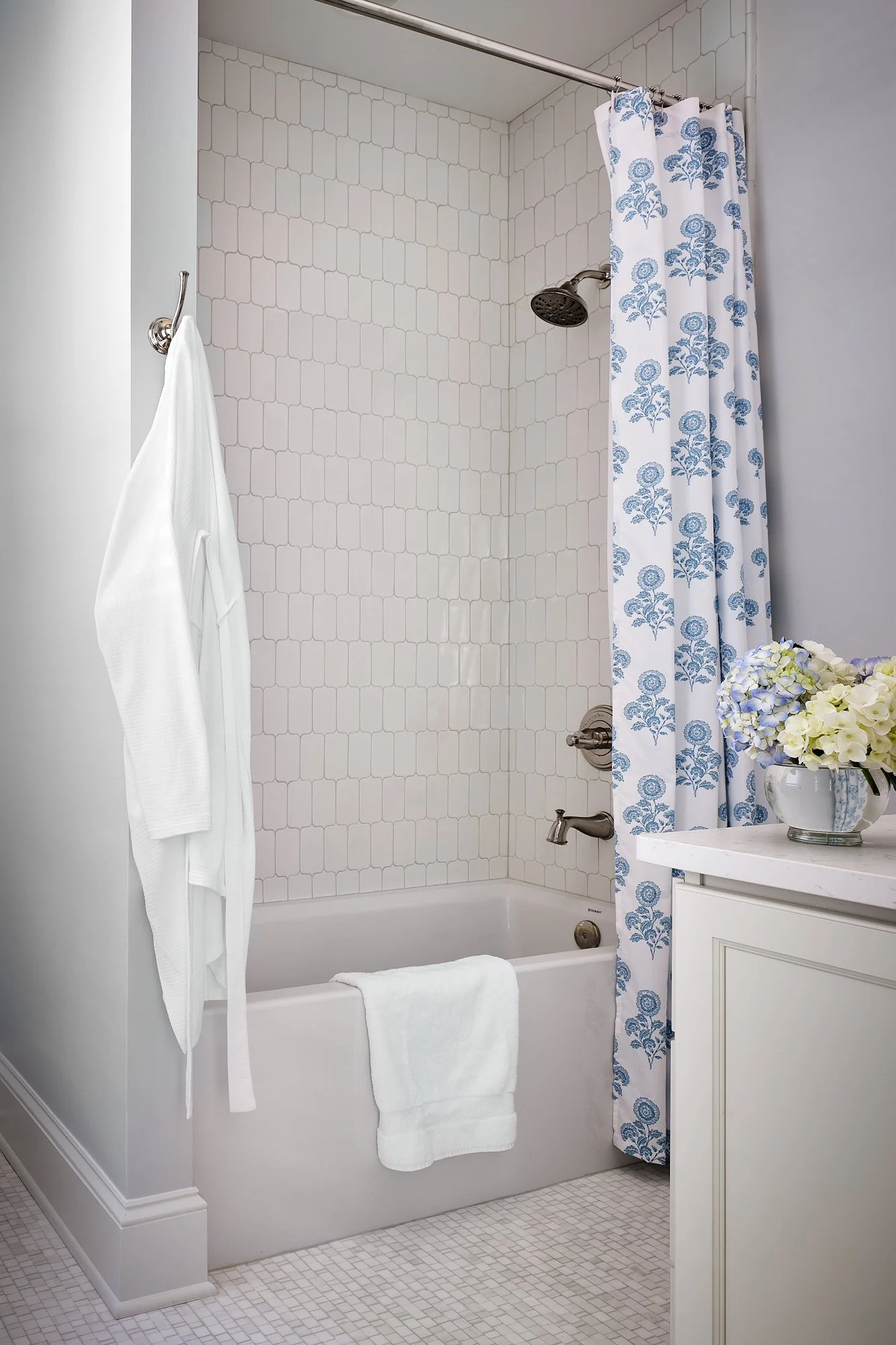 Bathroom with a bathtub and shower, white tiles, a shower curtain with blue floral print, a white towel on the bathtub, a white cabinet with a flower vase with white and purple flowers, and a towel hanging on a hook.