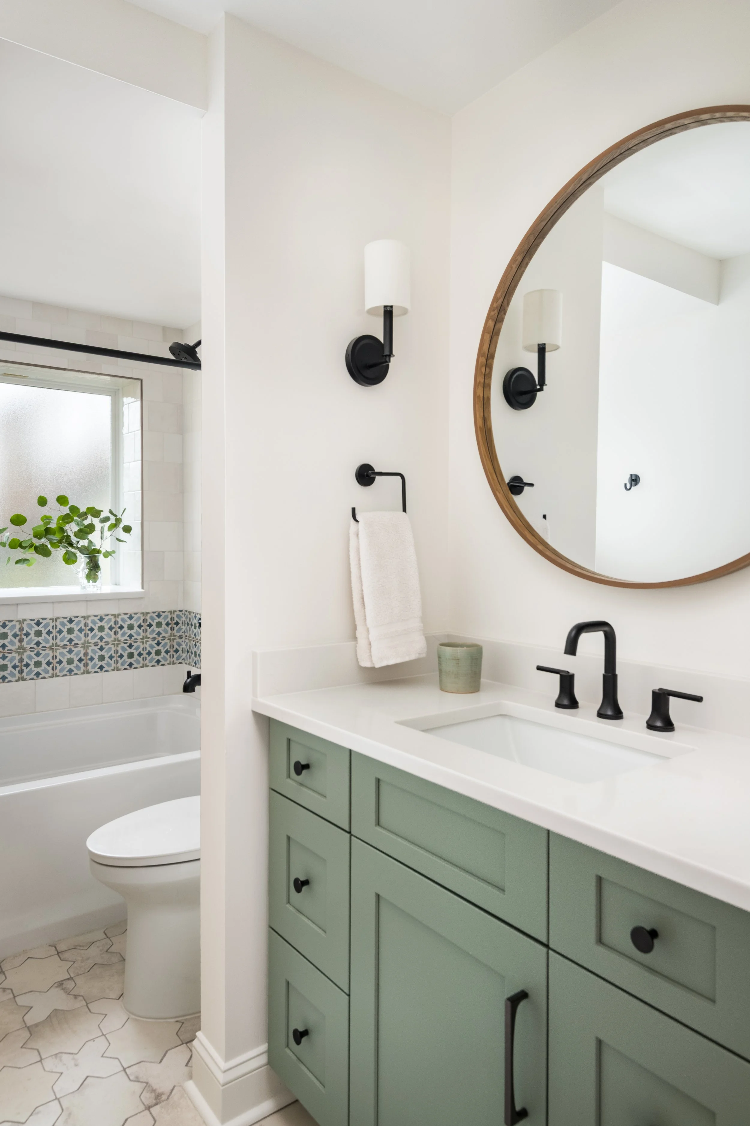 Bathroom with a green vanity, black hardware, a white countertop, a round mirror with wooden frame, a wall-mounted light, a towel ring with a towel, a small potted plant, a white sink, black faucet, and a bathtub with patterned tile border and window