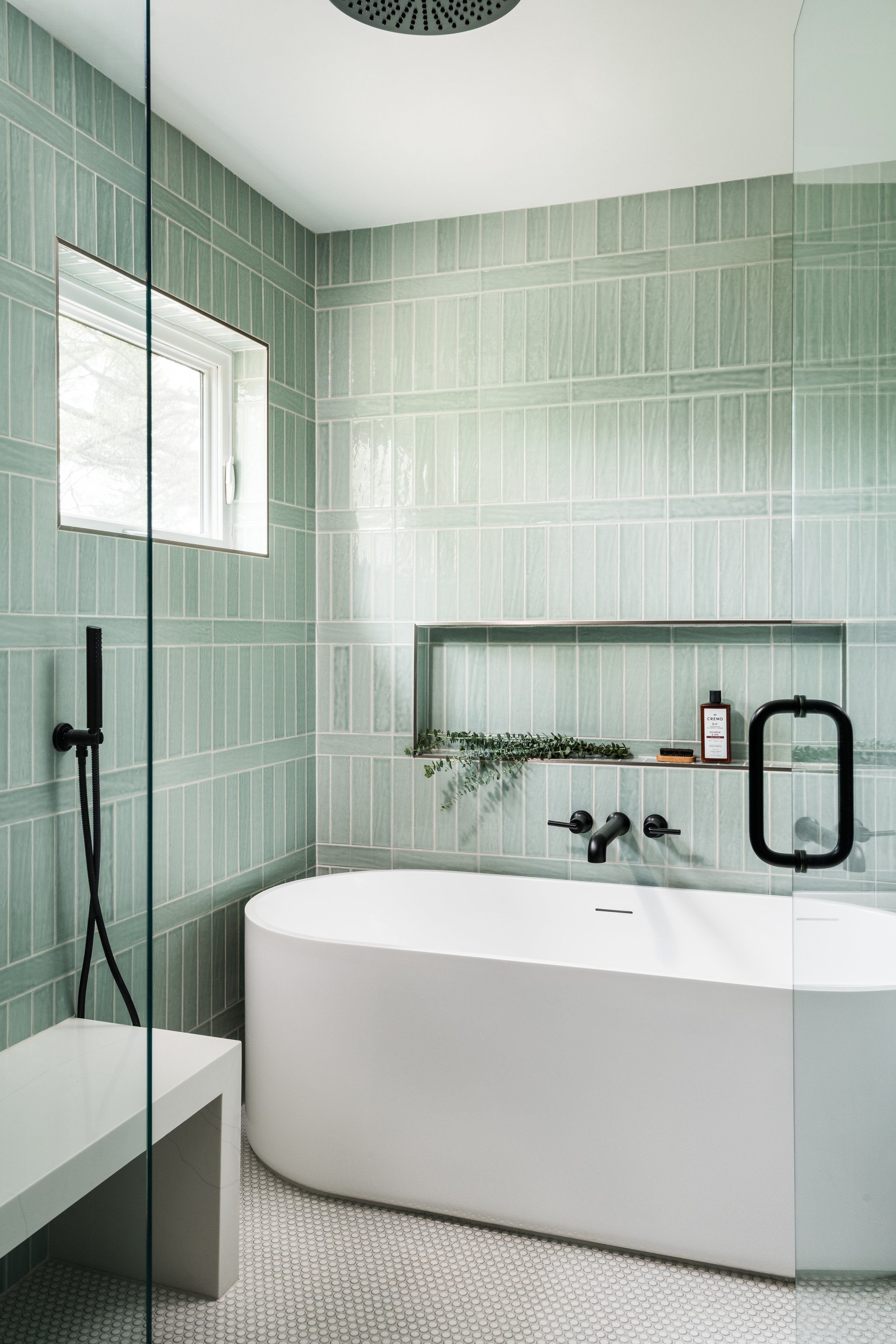 Modern bathroom with green tiled walls, a white curved bathtub, black fixtures, a small window, and a mirrored wall.