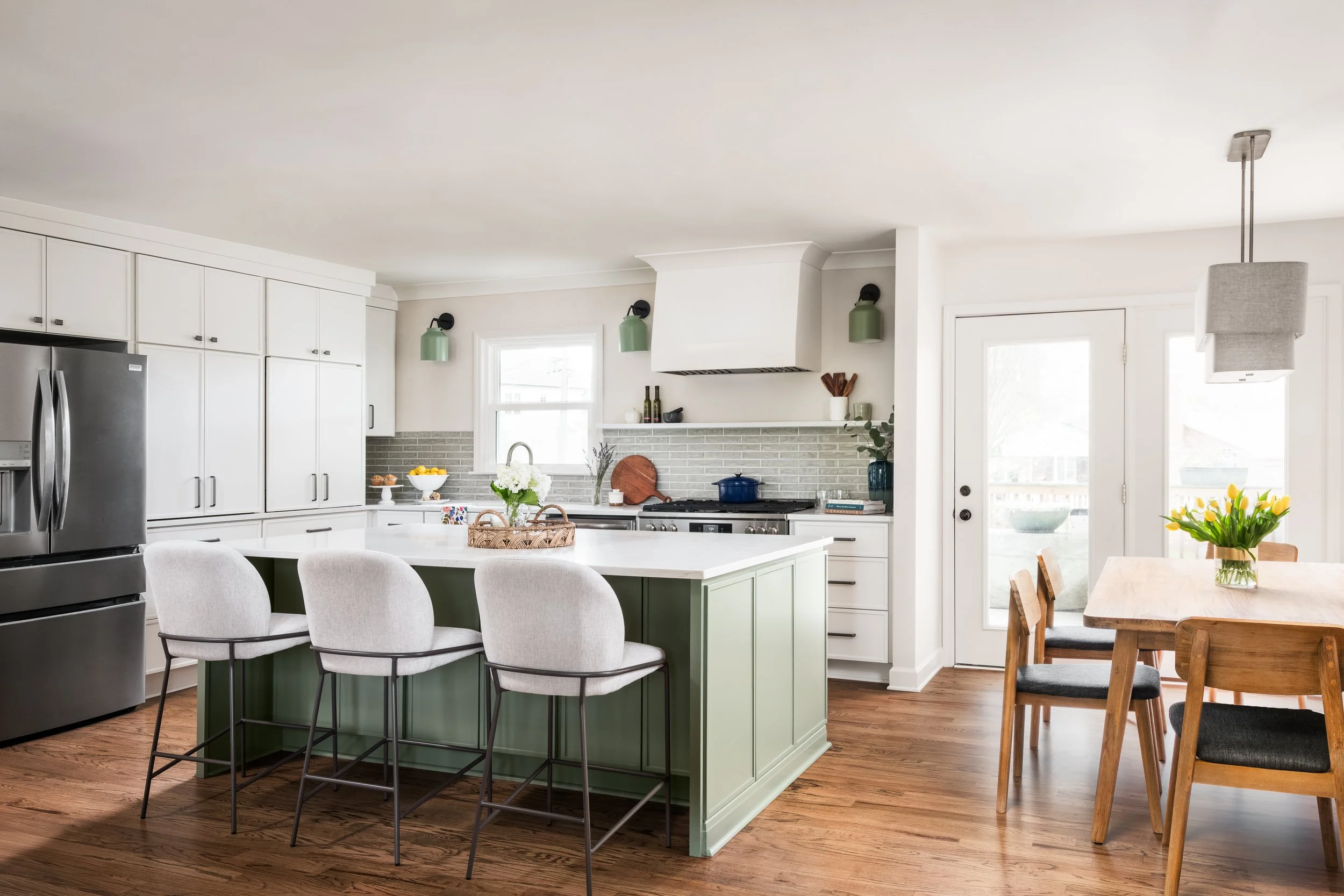 A modern kitchen with white cabinets, a green island, stainless steel refrigerator, and a dining table with chairs. There are decorative items including a flower vase on the island and a bouquet of yellow flowers on the dining table.