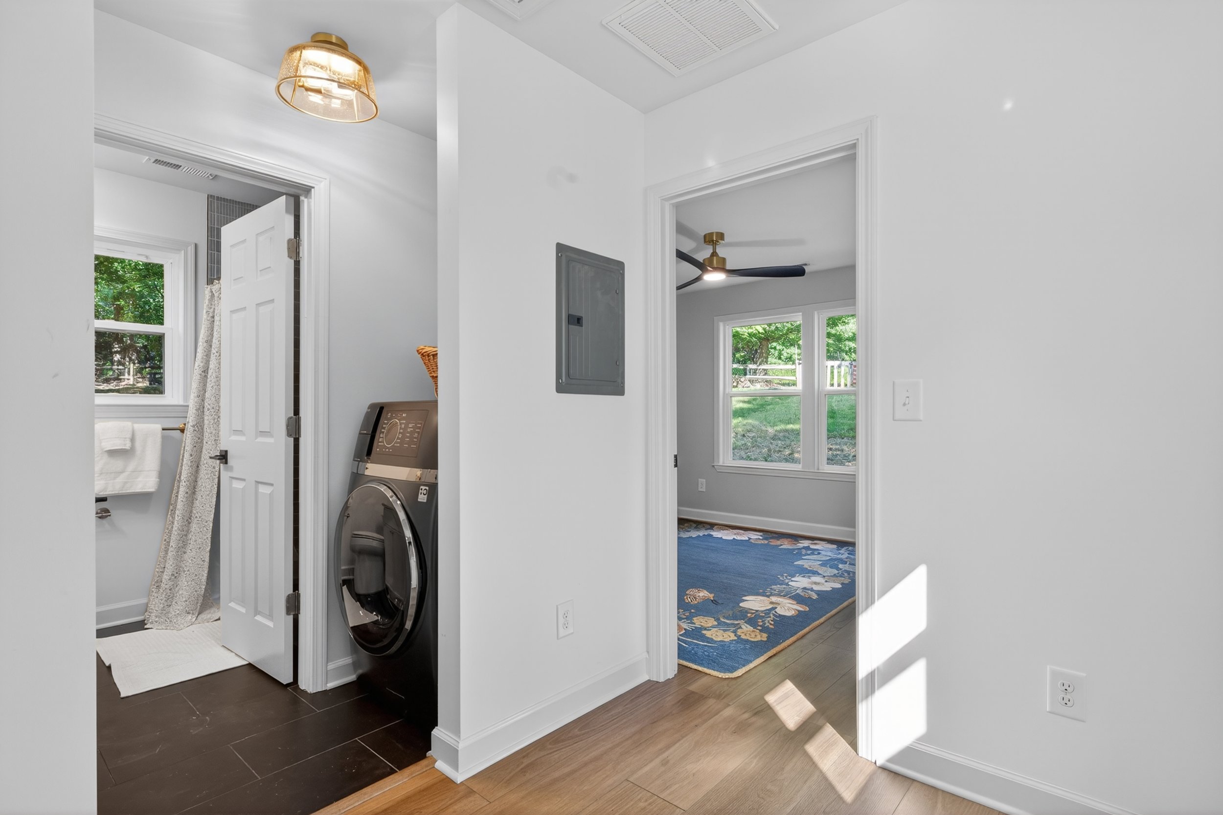 Inside view of a house showing a laundry area with a washing machine, an open door to a bathroom with a window and a towel, and a hallway with carpet and windows. The ceiling has light fixtures and ventilation diffusers.