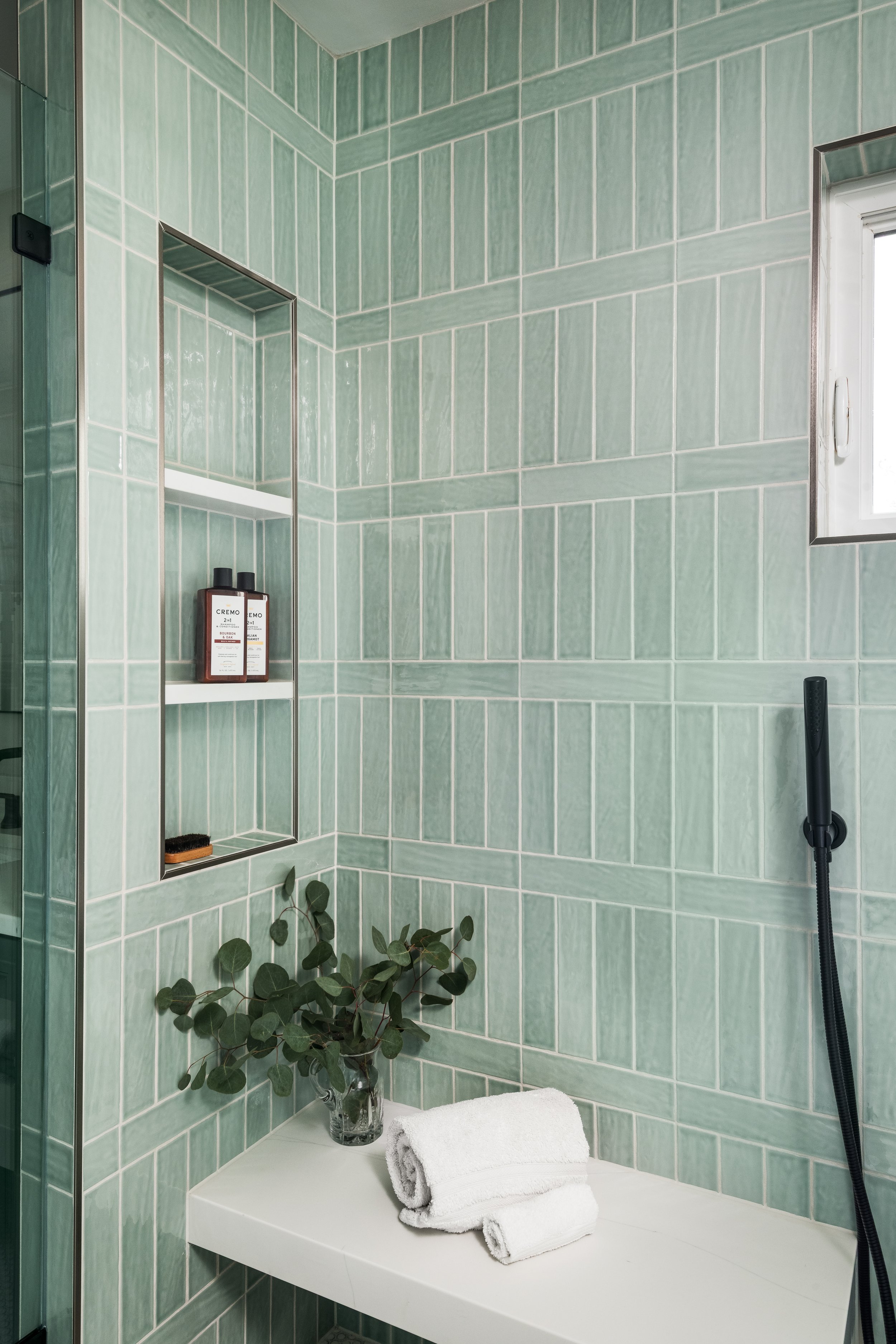 A modern bathroom with green tiled walls, a window, a built-in shelf with toiletries, a white countertop with rolled white towels, a glass vase with eucalyptus, and a black handheld shower head.