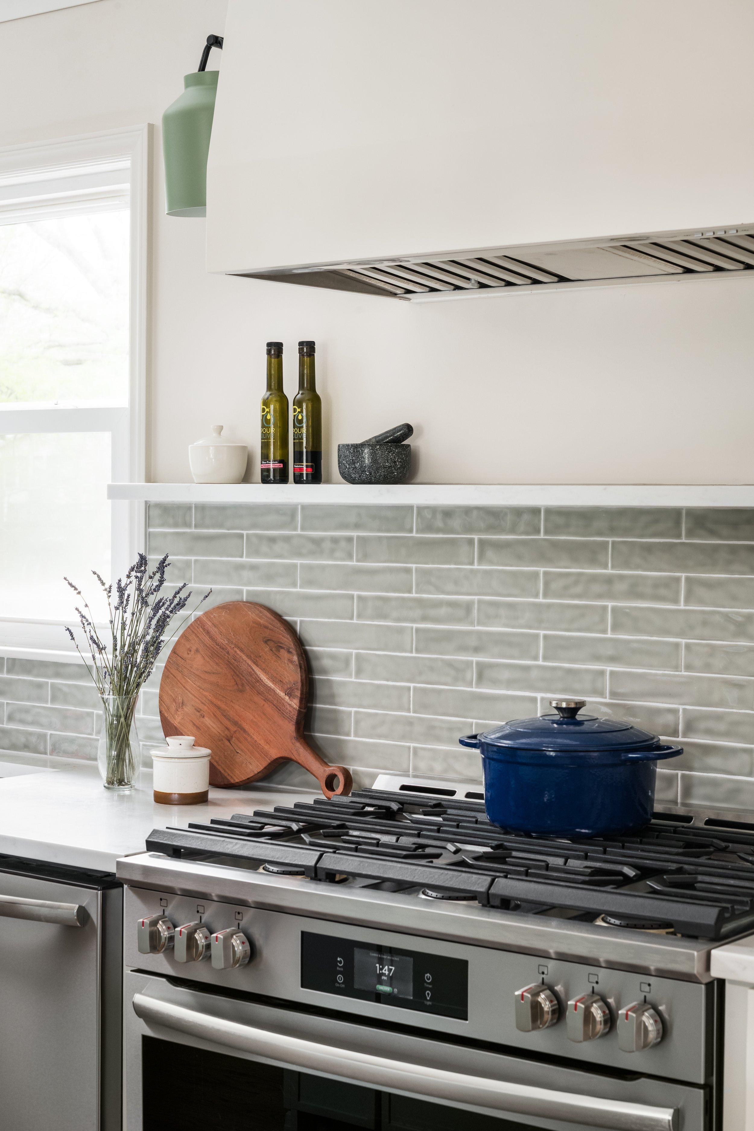 Modern kitchen with a stainless steel oven and stove, gray backsplash tiles, a dark blue pot on the stove, and a wooden cutting board leaning against the wall. There is a glass vase with lavender on the counter, two bottles of olive oil, a mortar and