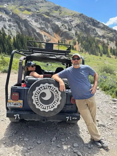 Two people, a man and a woman, are outdoors near a black off-road vehicle with a spare tire cover. The man is standing beside the vehicle, leaning on it with his right arm, wearing a blue t-shirt, pants, sunglasses, and a cap. The woman is sitting inside the vehicle, wearing sunglasses and a hat. The background features a mountainous landscape with green grass, rocky slopes, and a clear blue sky.