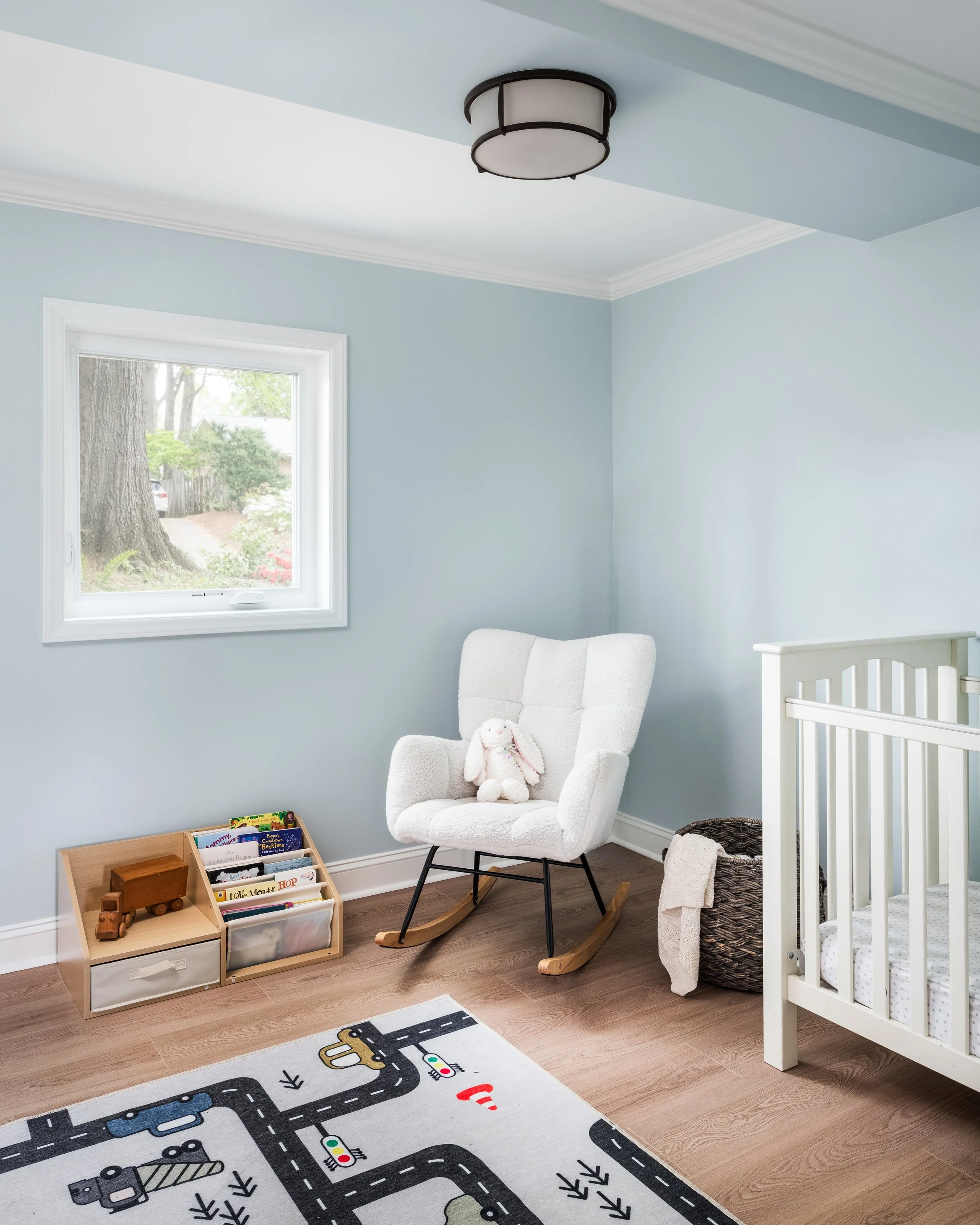 A corner of a nursery room with a white crib on the right, a white armchair with a stuffed bunny, a rocking chair, a window with trees outside, a wooden bookshelf with children's books, a toy truck, and a road-themed rug on the wooden floor.