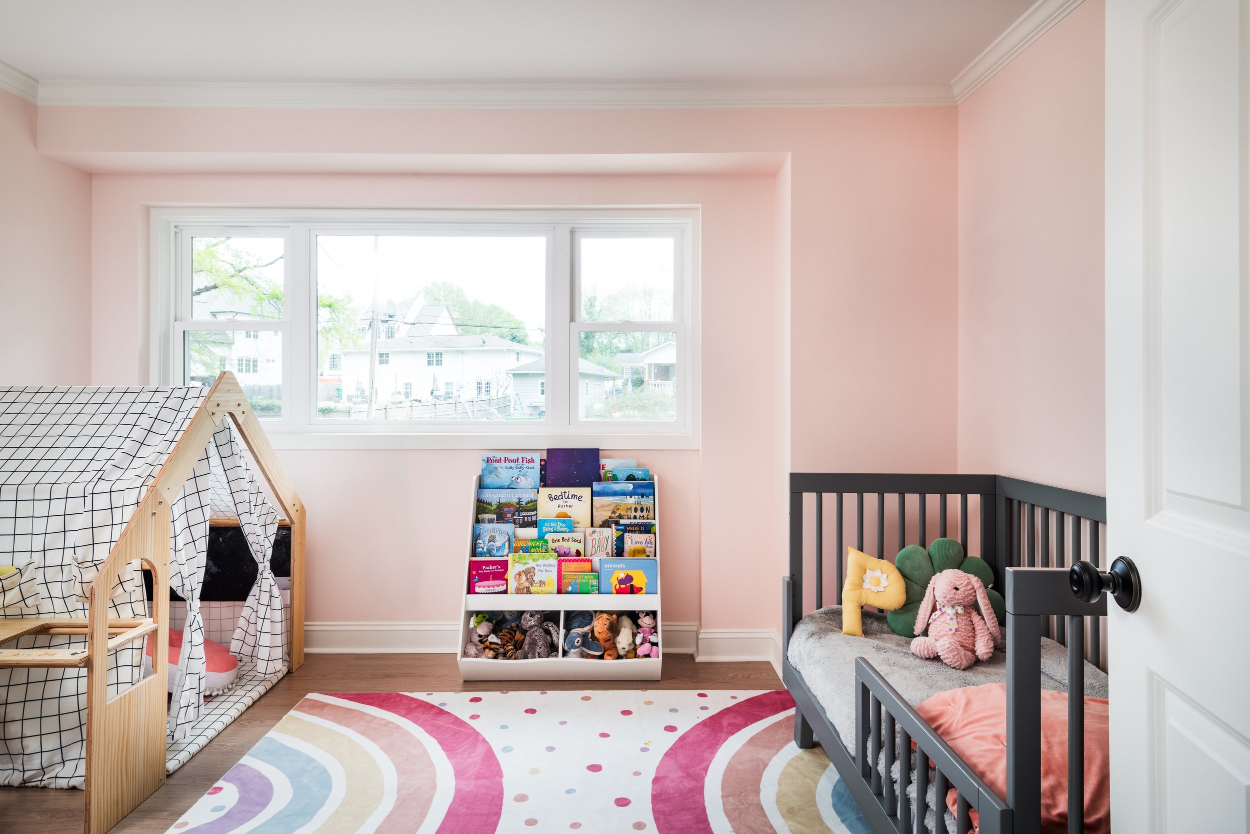 A colorful children's bedroom with a black crib, a stuffed pink bunny, various plush toys, a book display, and a rainbow-patterned rug. The room has large windows and pink walls.