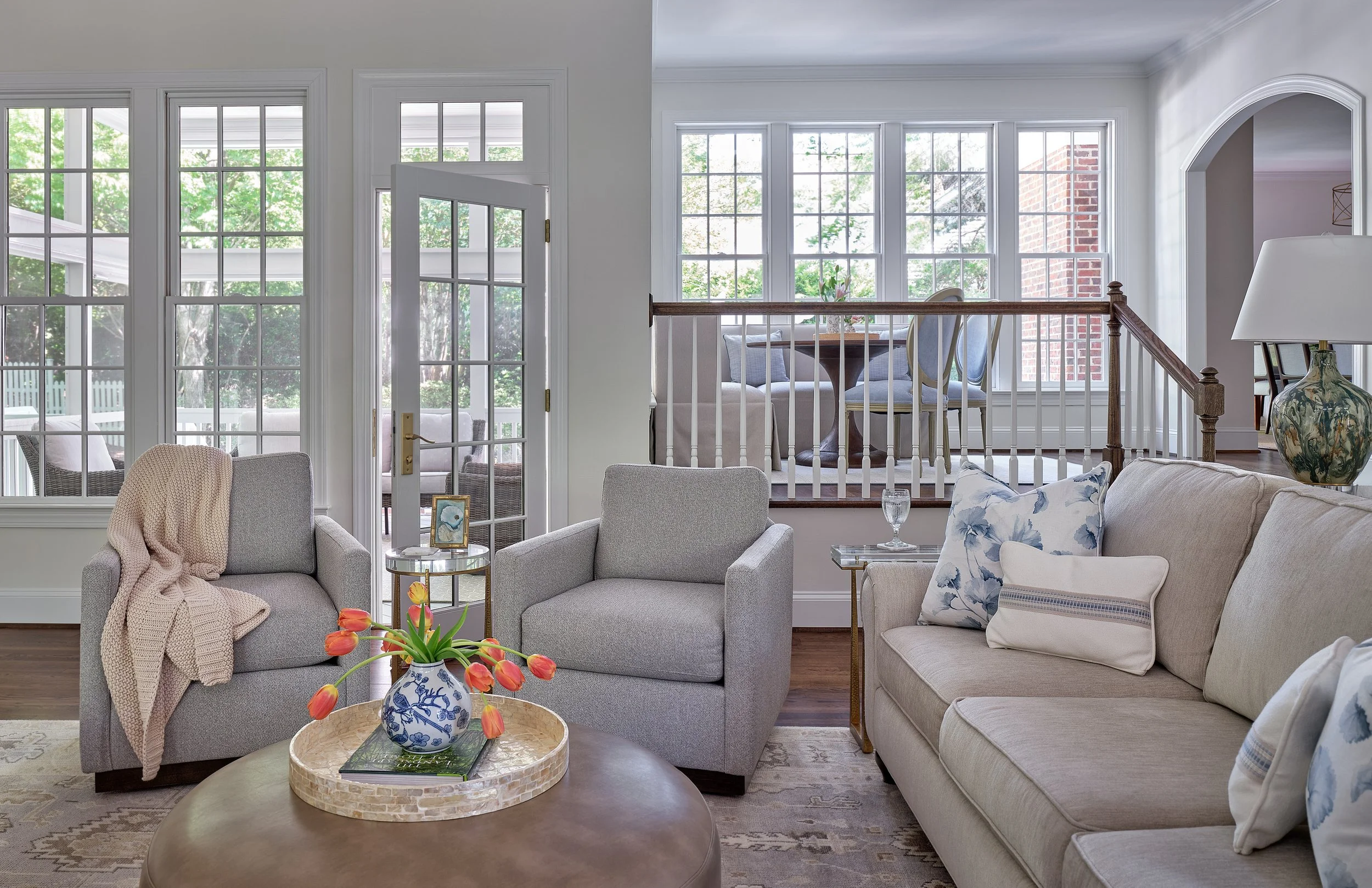 Living room interior with large windows, gray sofas, a round coffee table with a decorative blue and white vase of orange tulips, and a second story balcony overlooking the space.