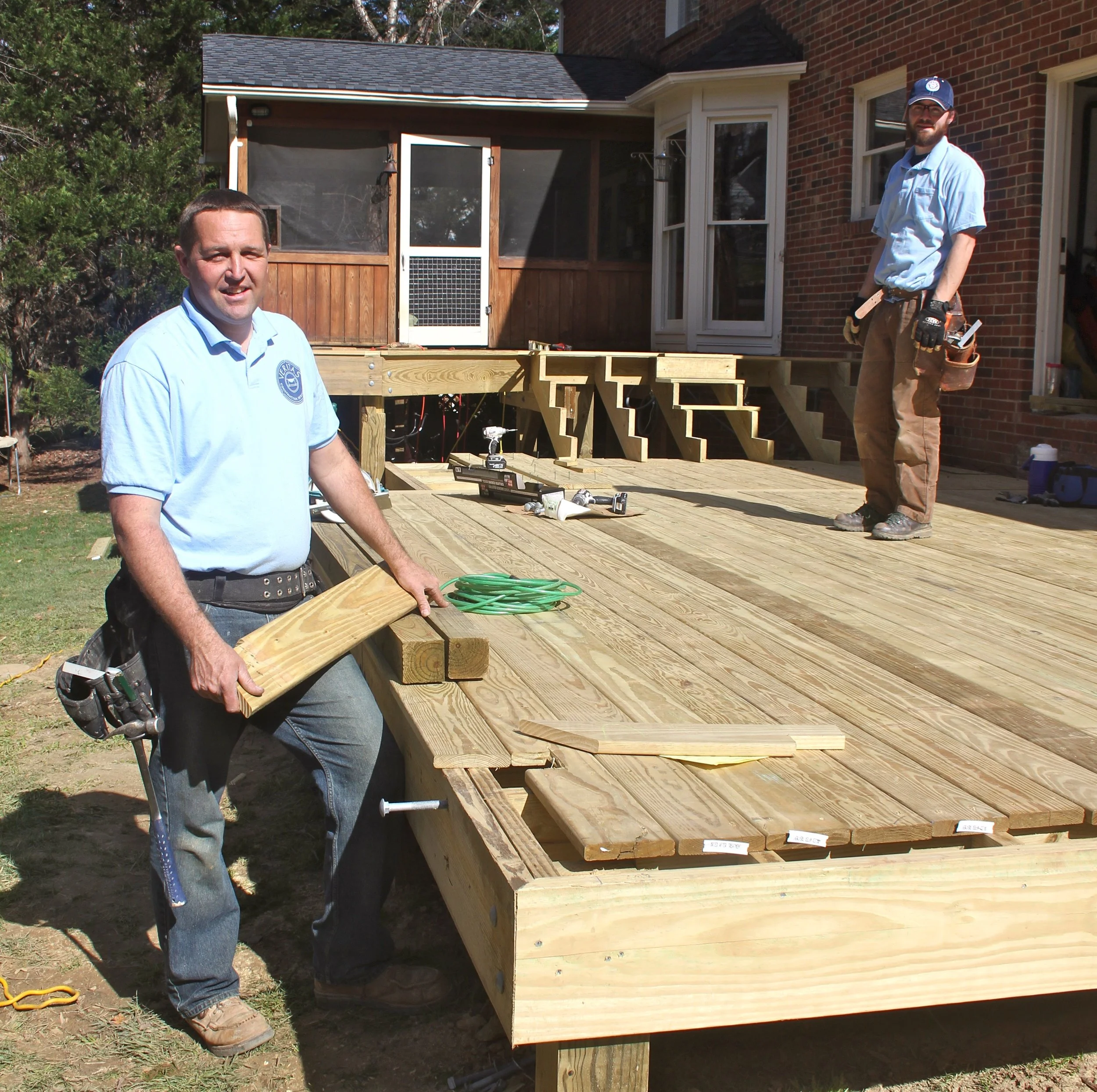 Two men building a wooden deck in the backyard of a brick house. One is wearing a light blue shirt and the other is wearing gloves and a cap. Construction tools are on the deck.