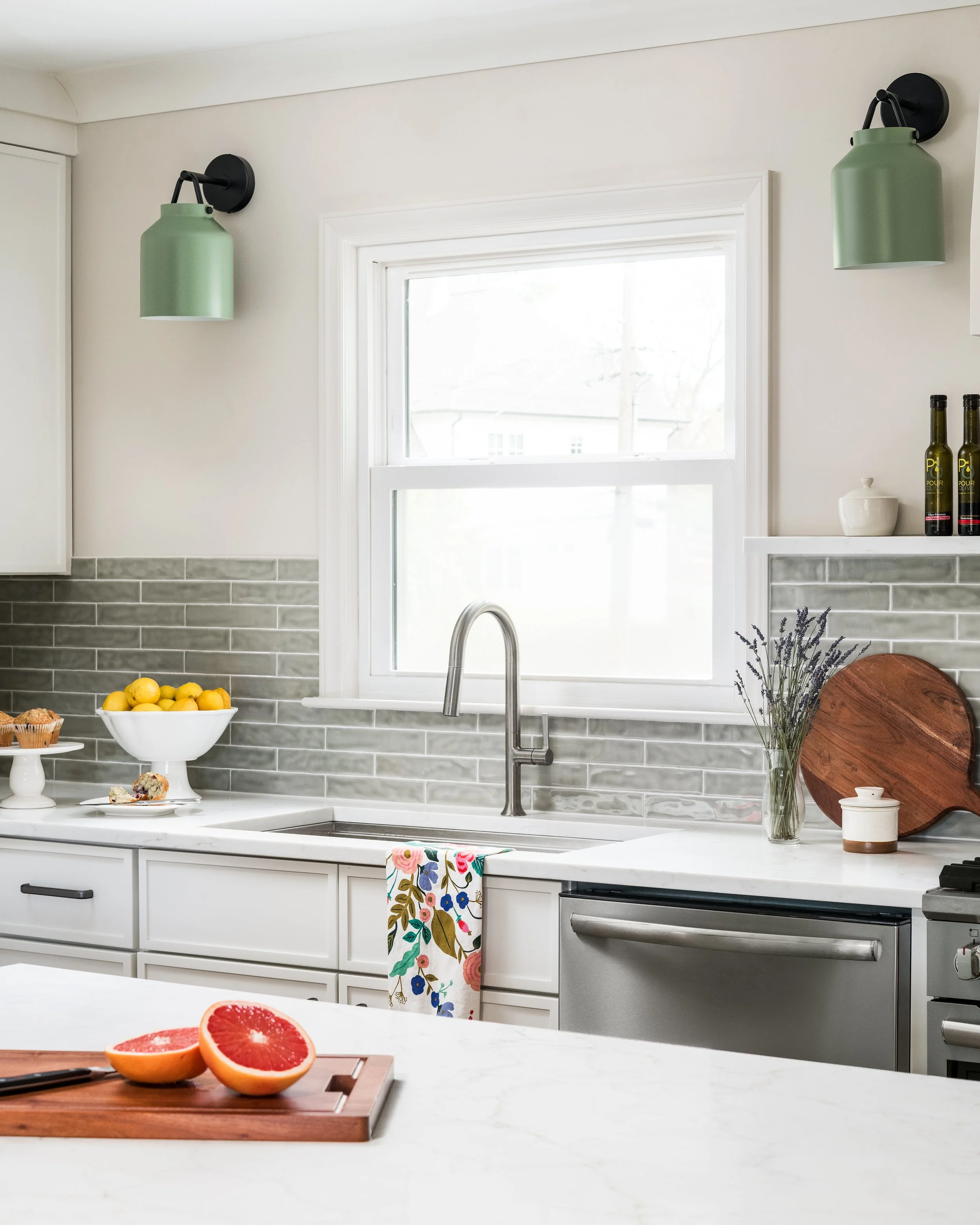 A modern kitchen with a white marble countertop, a stainless steel sink, and white cabinets. Two green wall-mounted lamps are on either side of a window. A bowl of lemons and a small cake on a stand are on the counter, with a colorful floral towel ha
