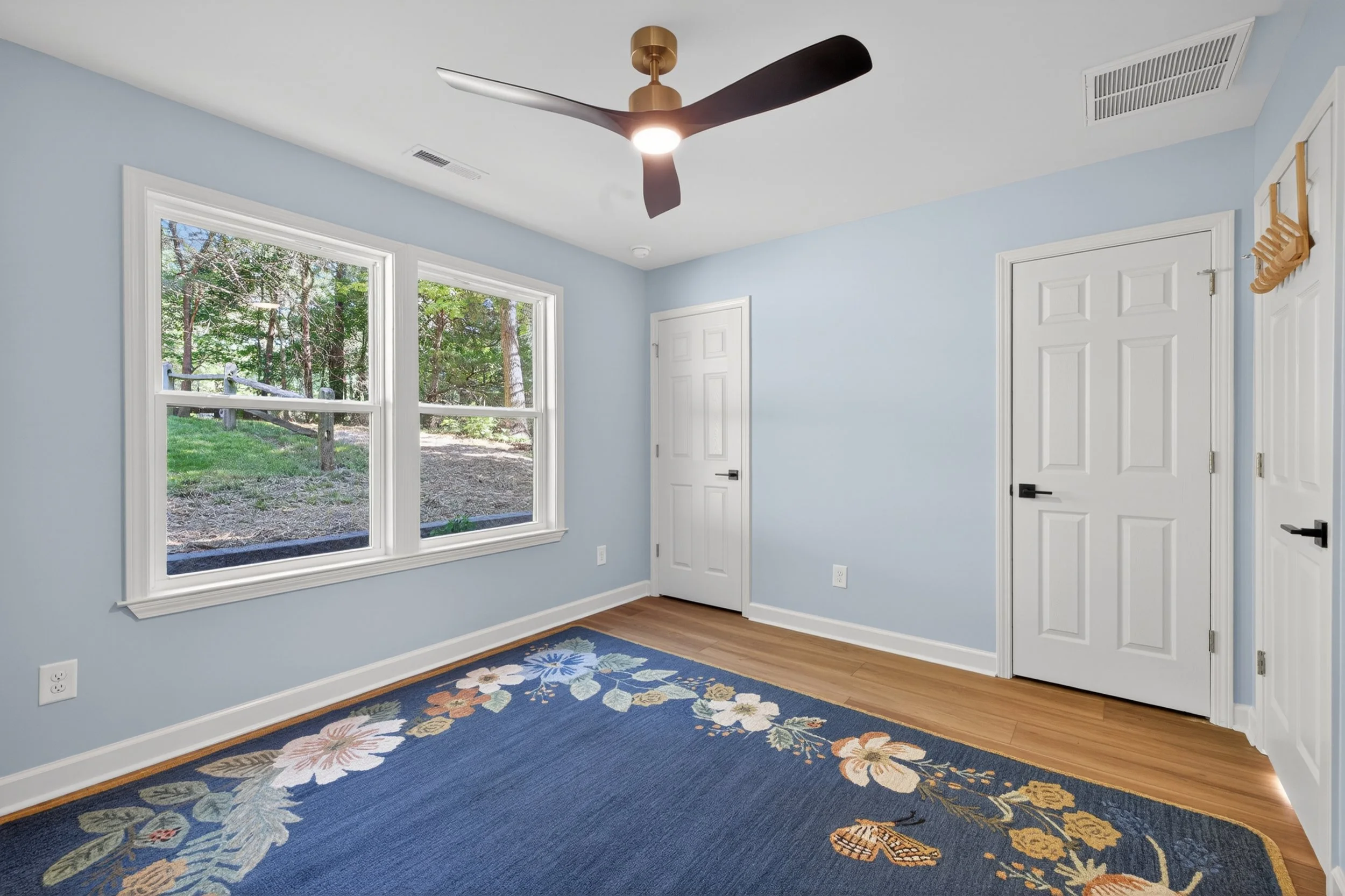 Interior of a bright room with blue walls, large windows showing trees outside, a ceiling fan, a door, a floral rug, and a small coat rack.