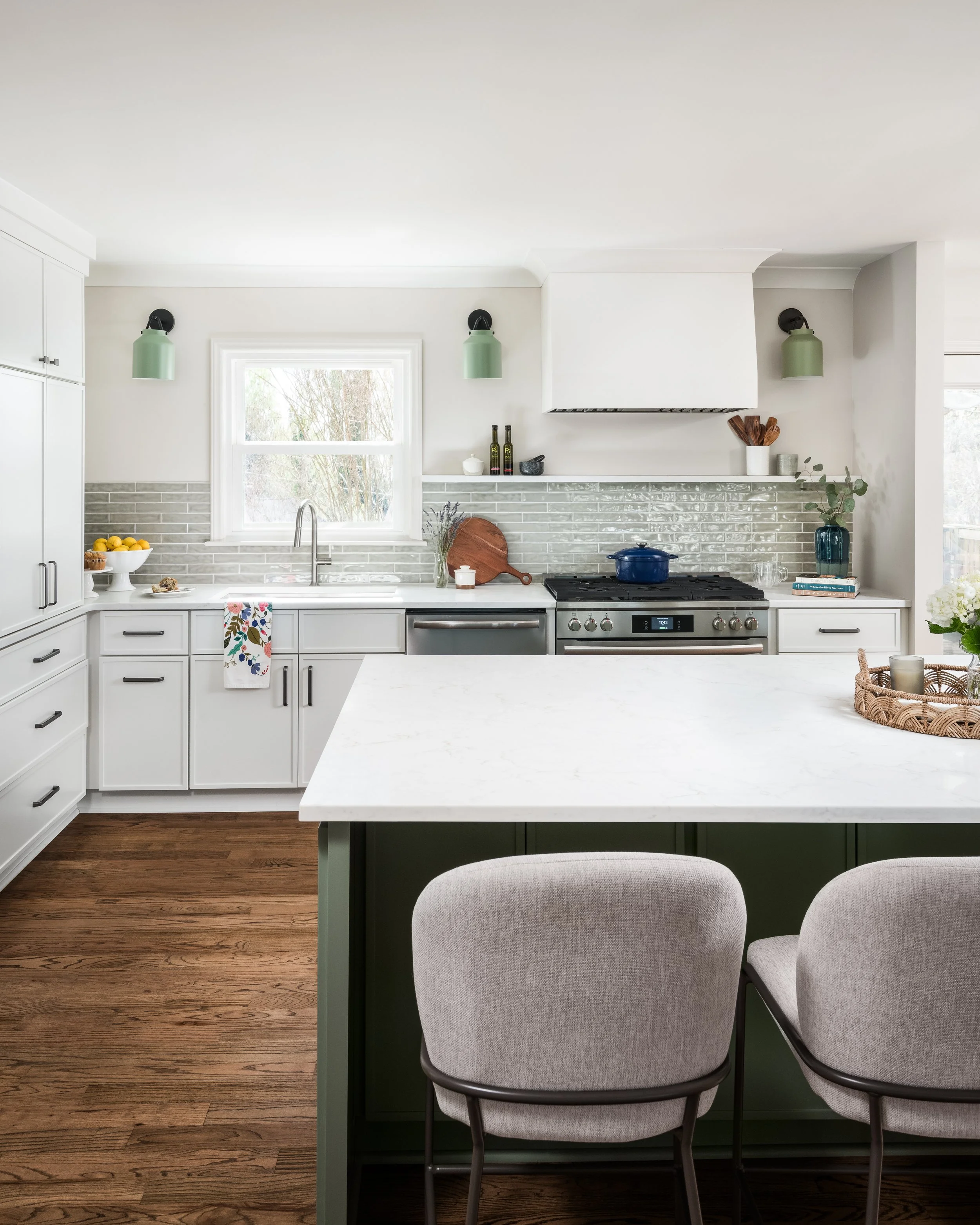 Modern kitchen with white cabinets, gray backsplash, and stainless steel oven. Green pendant lights and decorative items on the counter. Wooden floor, window with view of trees, and a marble kitchen island with beige upholstered chairs.