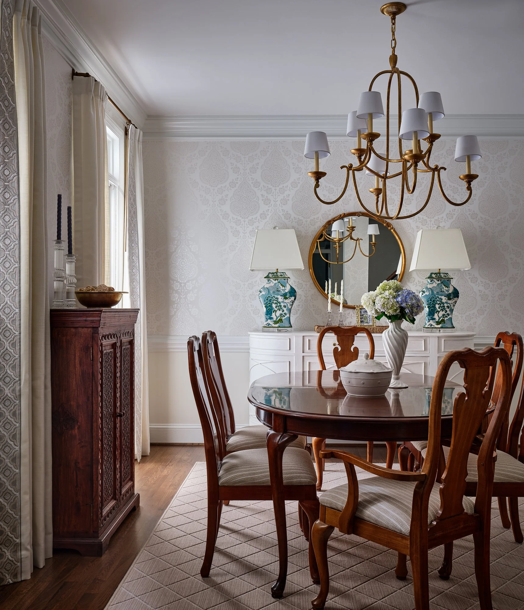 A dining room with a wooden table, eight chairs with beige upholstered seats, two blue and white vases, a white pitcher with flowers, and a chandelier hanging from the ceiling. The room has light-colored patterned wallpaper, a mirror, two lamps, and 