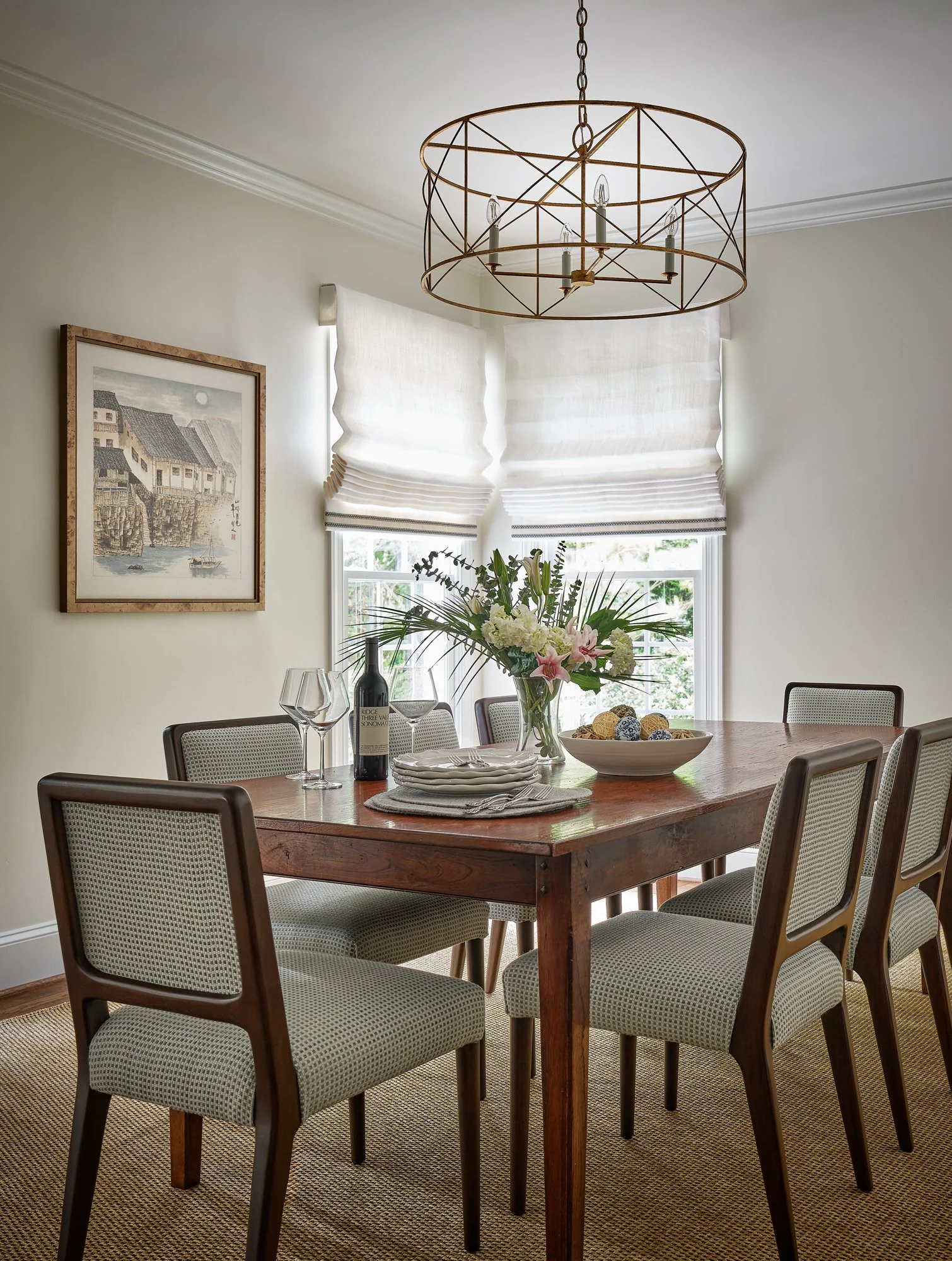 A dining room with a wooden table, six upholstered chairs, a vase with flowers, a bowl of decorative balls, wine glasses, a bottle of wine, a chandelier, and windows with white shades.