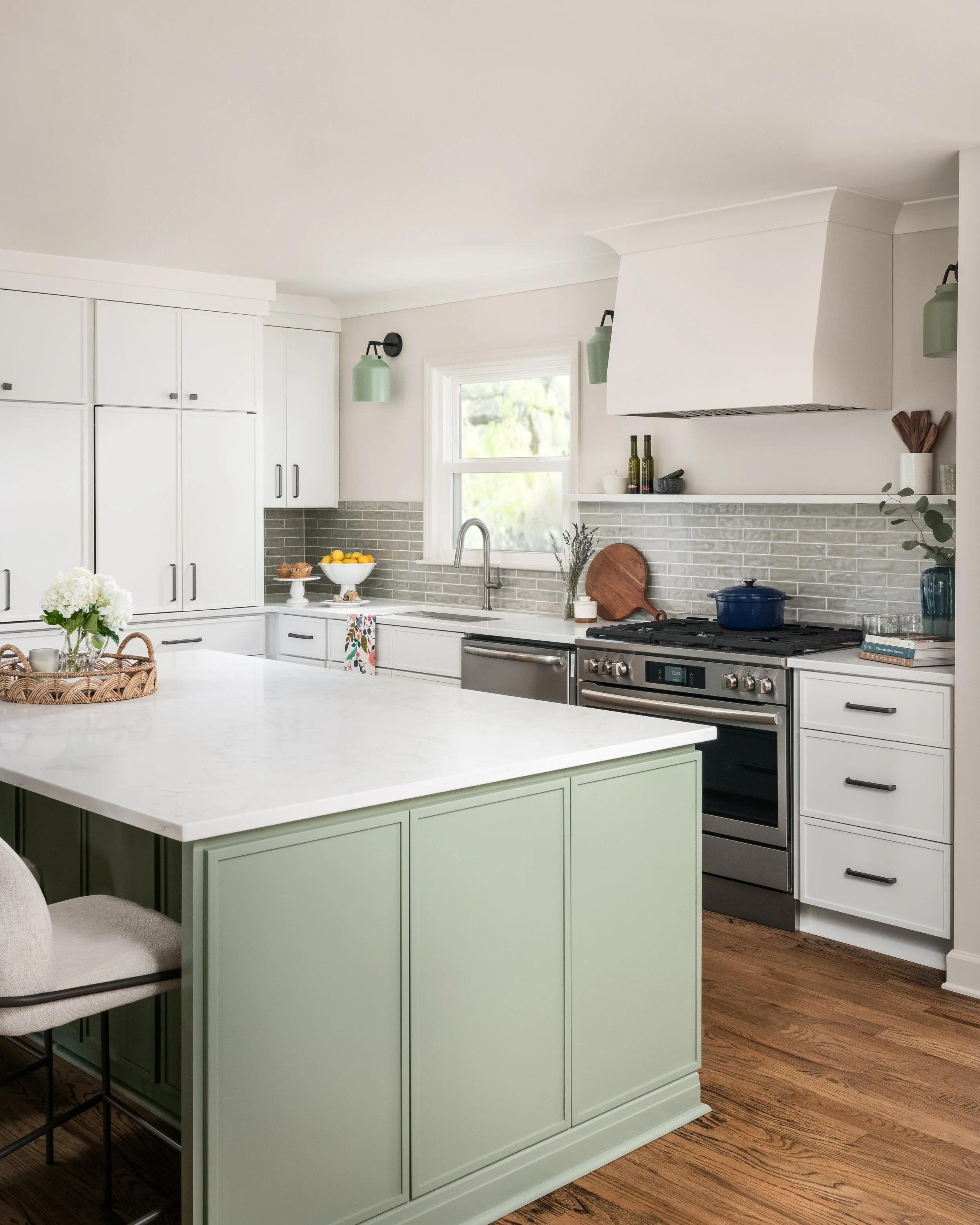 Modern kitchen with white cabinets, a white island with a light green base, stainless steel stove, gray backsplash tiles, decorative items on the counter, and wooden flooring.