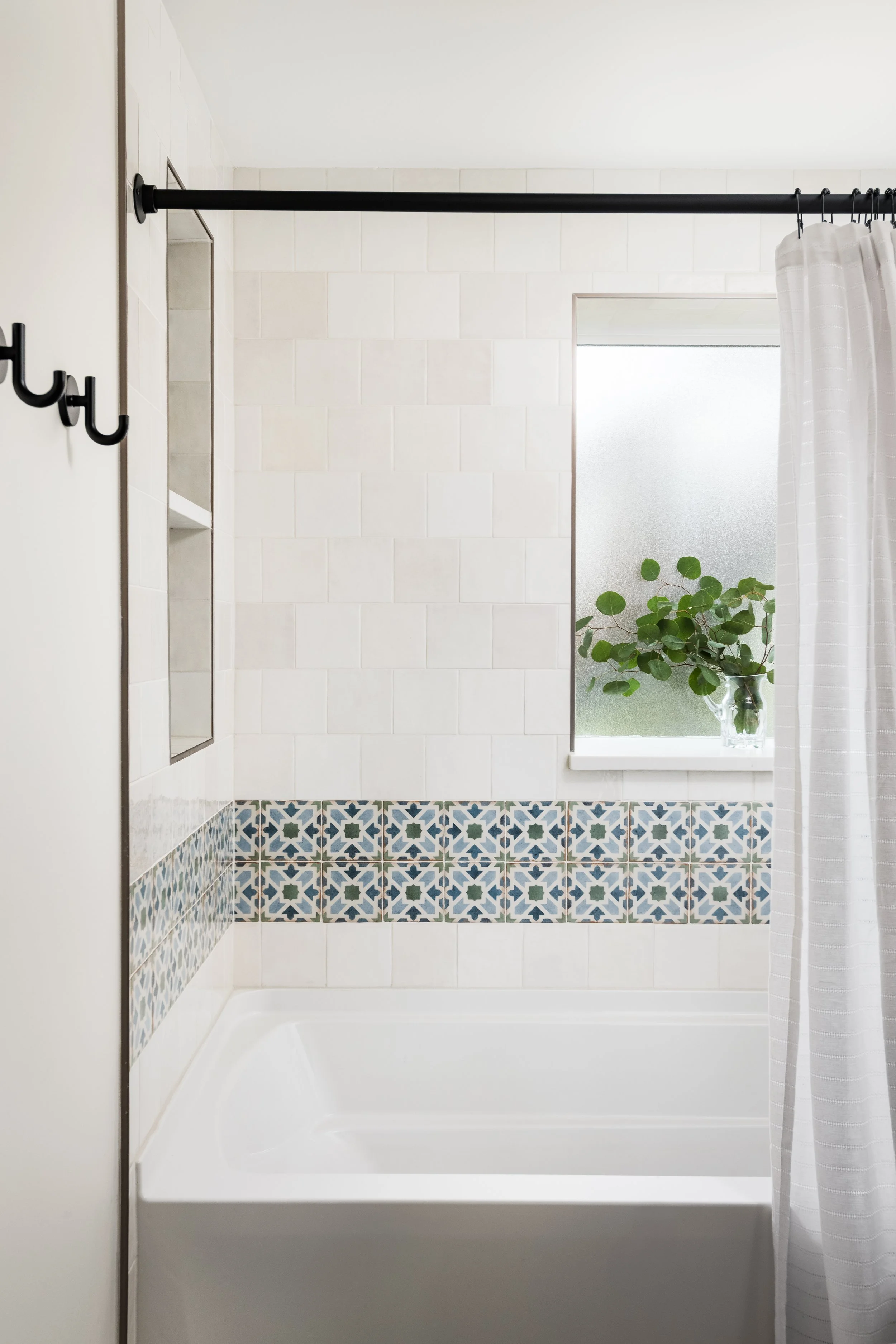 A white bathtub with a black curtain rod and white striped shower curtain. The wall behind has white tiles with a decorative border of blue, green, and white patterned tiles. A frosted window and a potted green plant are visible in the background.