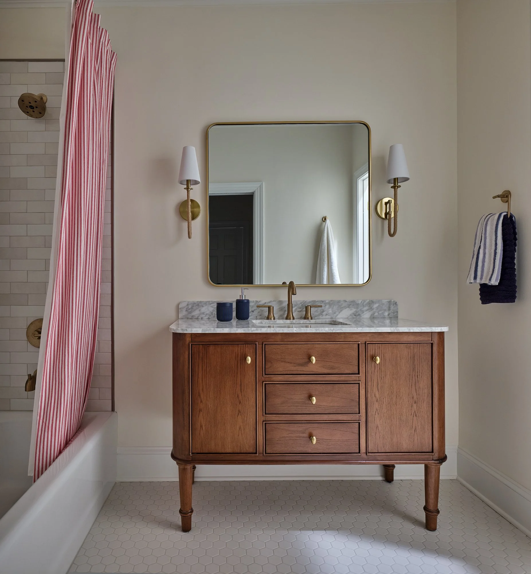 Bathroom vanity with marble top, wooden cabinet with brass knobs, wall-mounted lamps on either side, and a mirror above the sink. To the left, a shower with a striped red and white shower curtain, white subway tiles, and brass fixtures. Tiled white h
