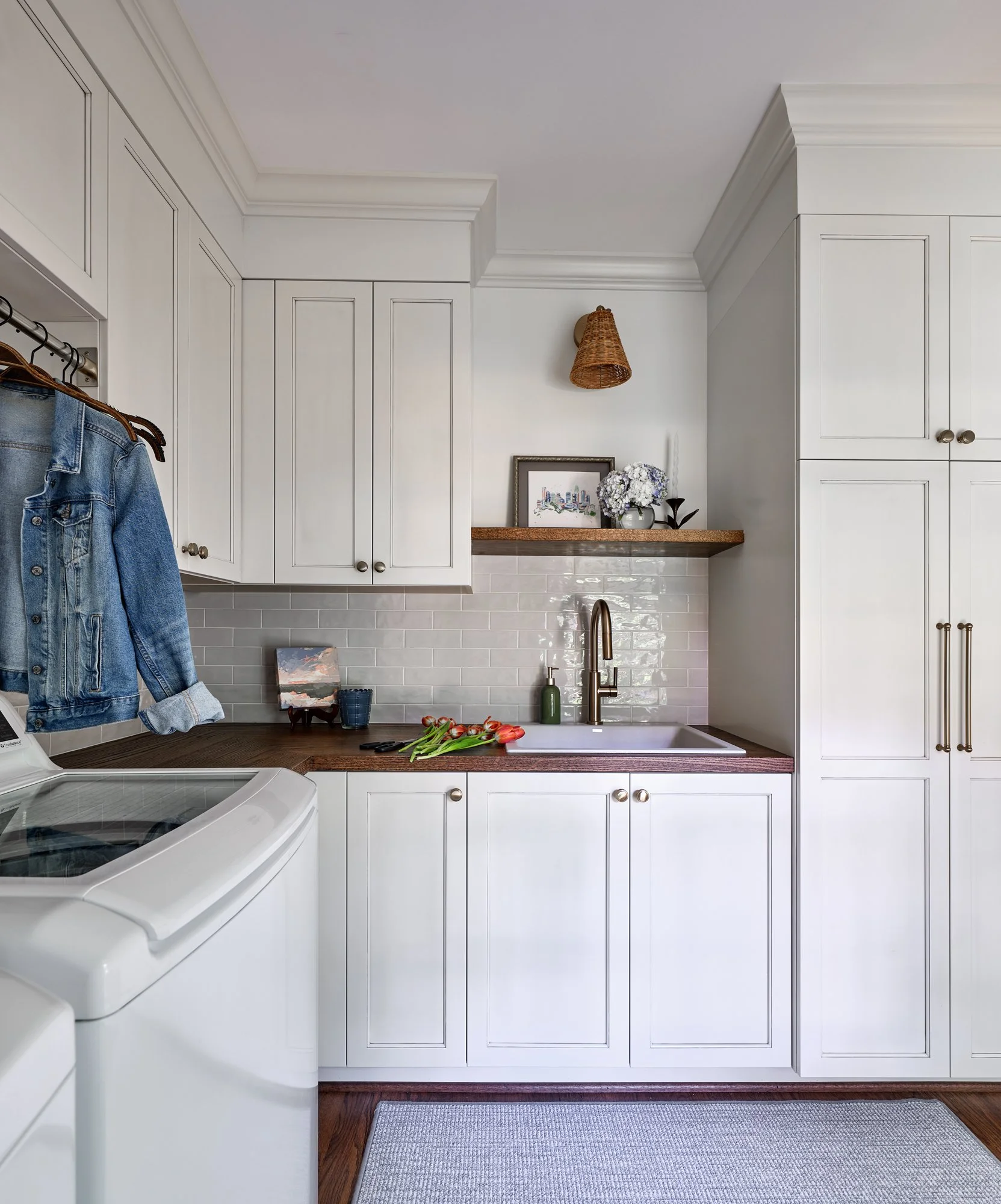A laundry room with white cabinets, a white tiled backsplash, a wooden countertop, and a white sink. On the countertop, there are flowers, a framed picture, and a plant. To the left, there's a washing machine with denim jackets hanging nearby. A smal